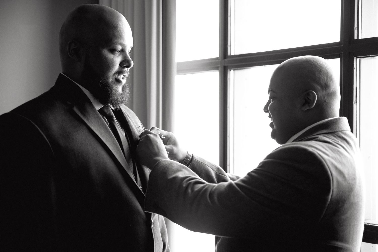 Black and white photo of two men adjusting a tie near a window, perfect for wedding preparation moments.