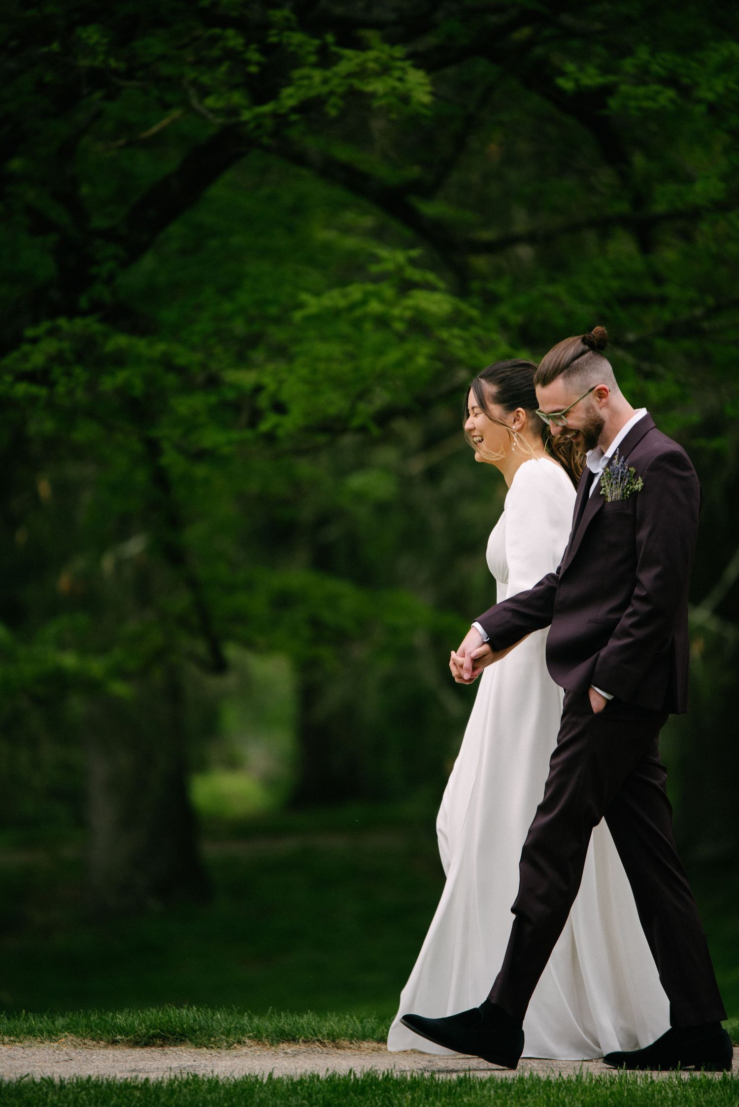 Bride and groom walk hand in hand through lush green park, smiling joyfully on their wedding day.