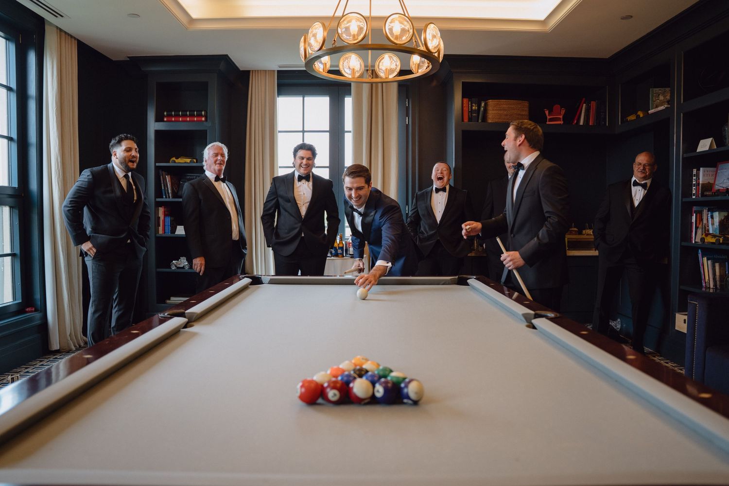 Groomsmen gather around a pool table in an elegant room as the groom breaks the rack during pre-wedding celebrations.