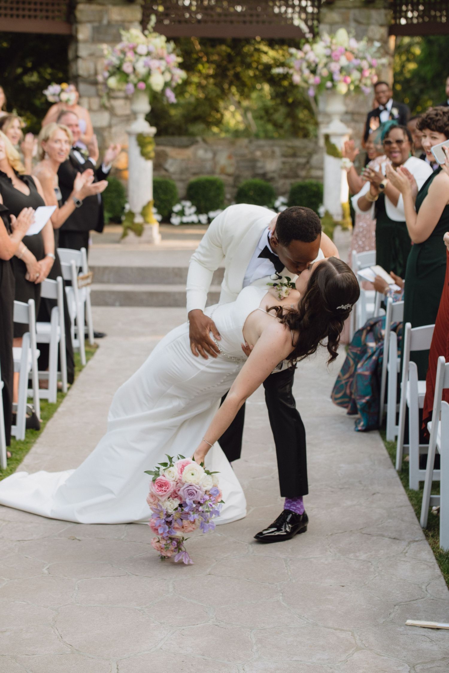 Bride and groom share a romantic dip kiss while walking down the aisle at an outdoor wedding ceremony.