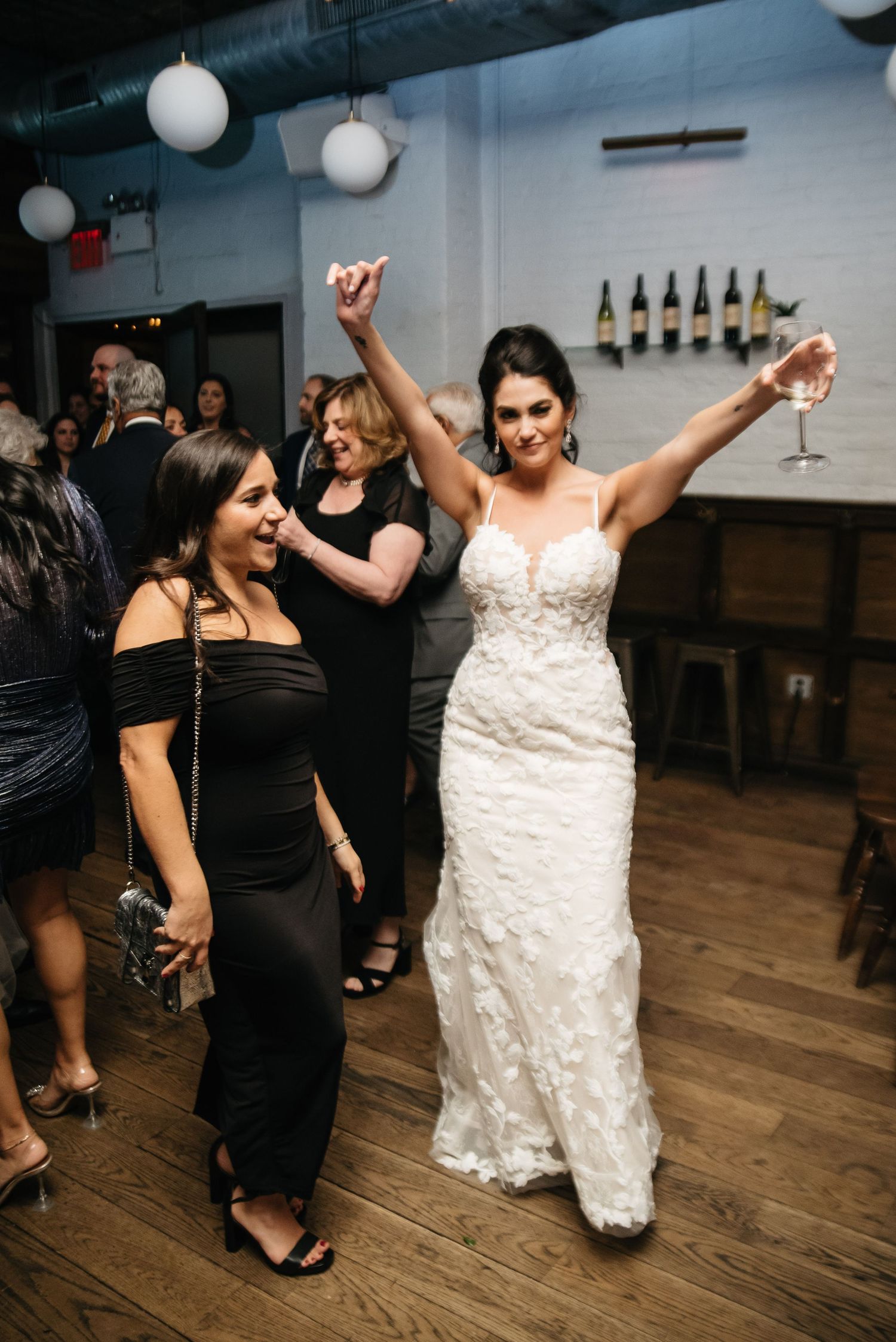 Bride in white lace gown raises arms joyfully on wedding reception dance floor while guests celebrate around her.