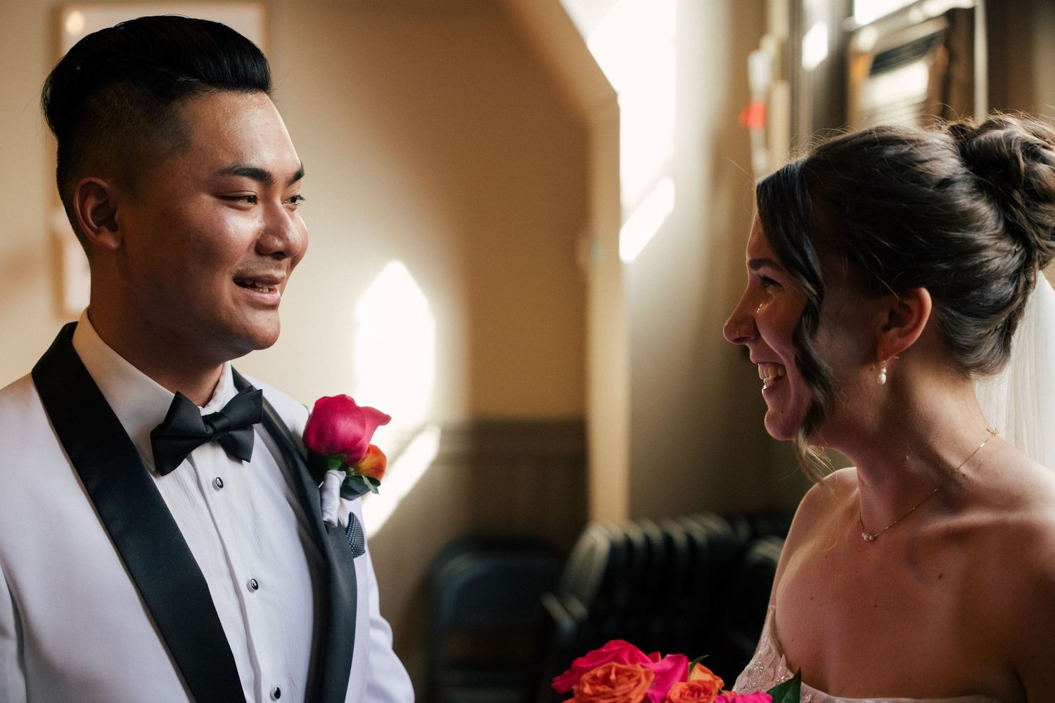 Bride and groom share a joyful moment, groom in black tuxedo with pink boutonniere, bride holding vibrant bouquet.