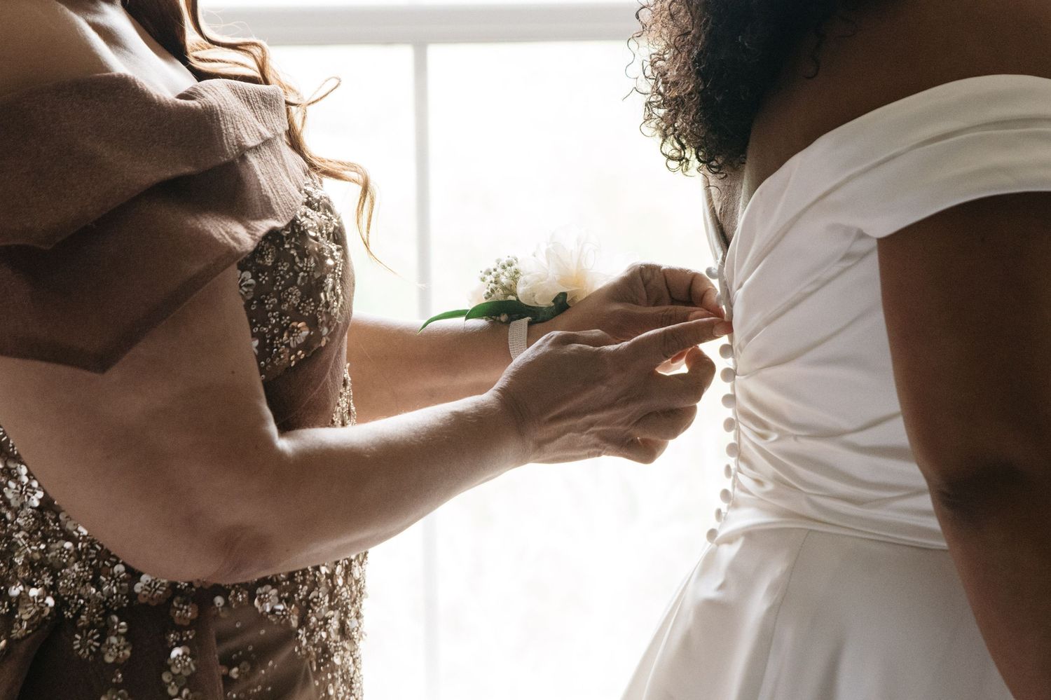 Bridesmaid helping bride button up white wedding dress near a bright window.