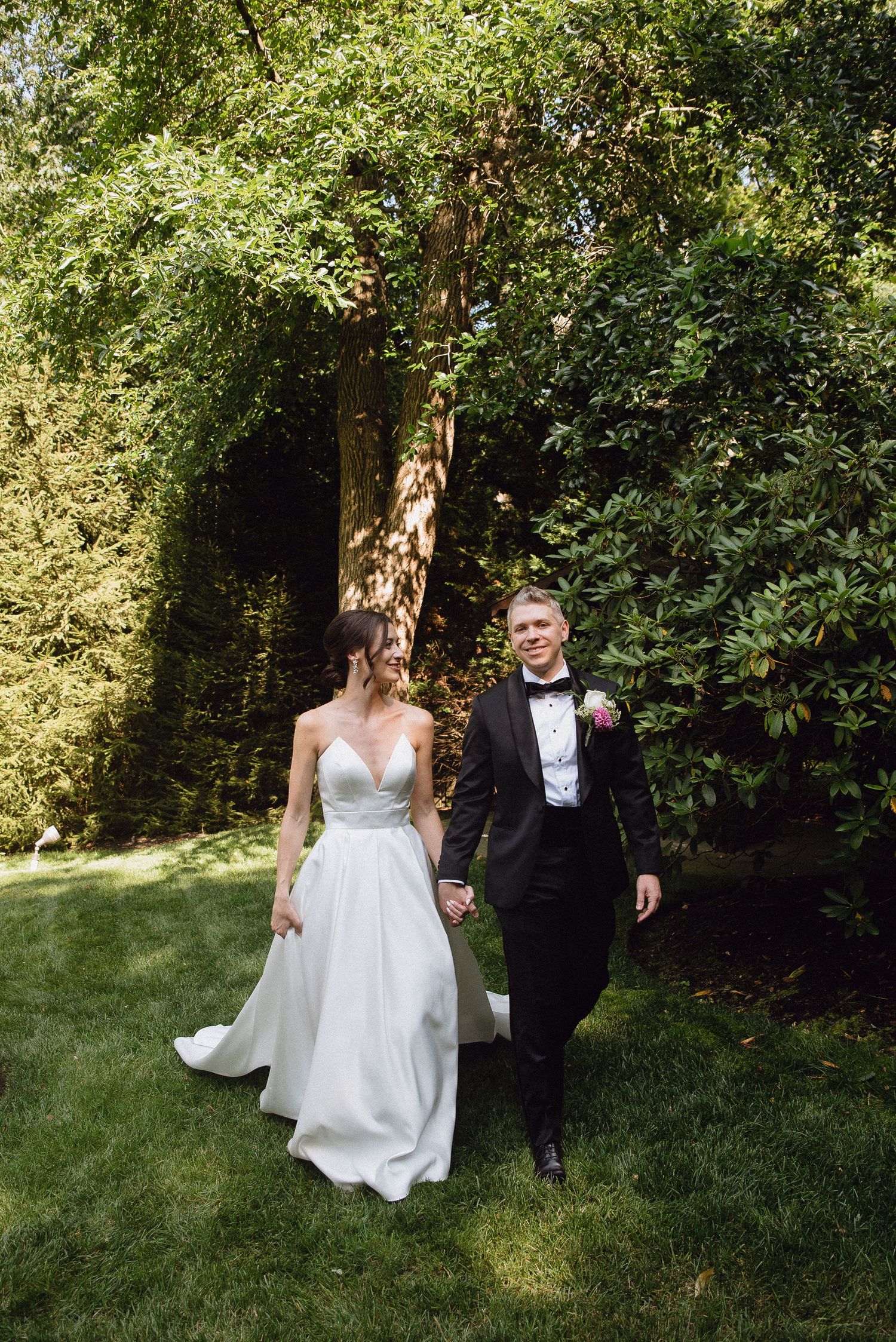 A bride and groom walk hand-in-hand through a lush green garden, she in a white gown, he in a black tuxedo.