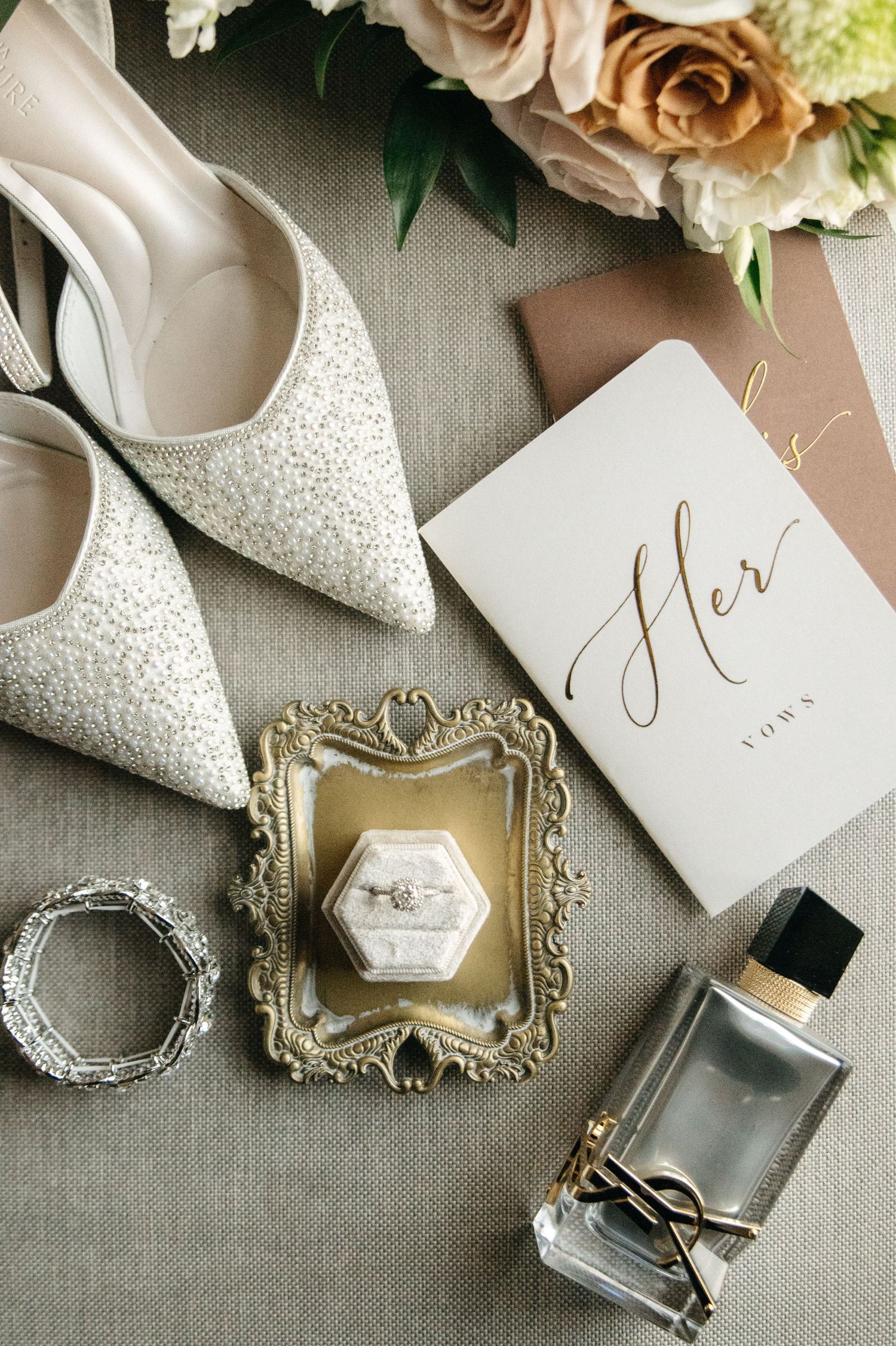 Elegant bridal flat lay featuring sparkly heels, diamond ring in velvet box, silver bracelet, perfume, vows book, and roses.