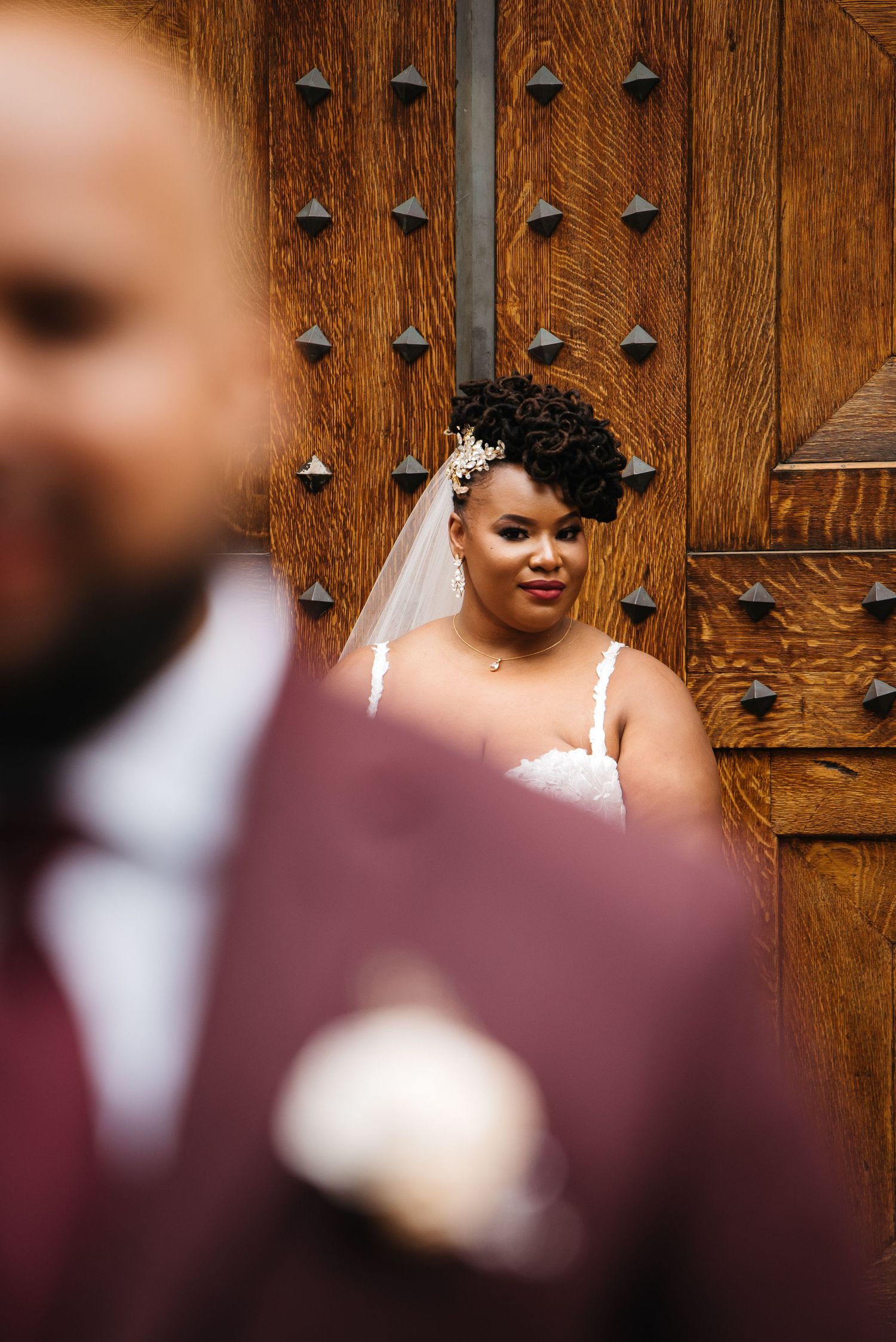 Bride in white lace gown and veil stands before ornate wooden door as groom in burgundy suit stands in foreground.