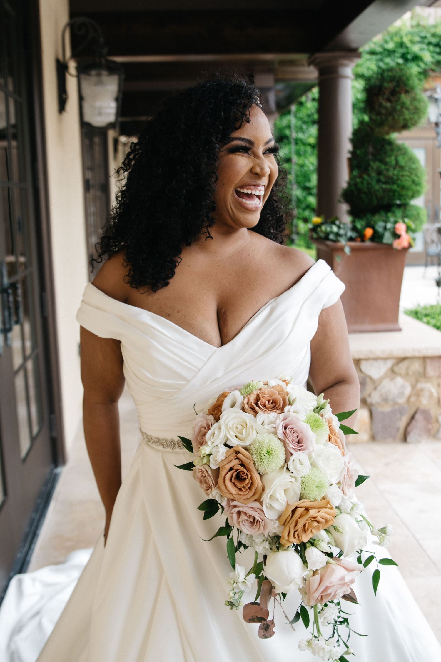 Joyful bride in off-shoulder white gown laughing while holding cascading bouquet of roses outdoors.