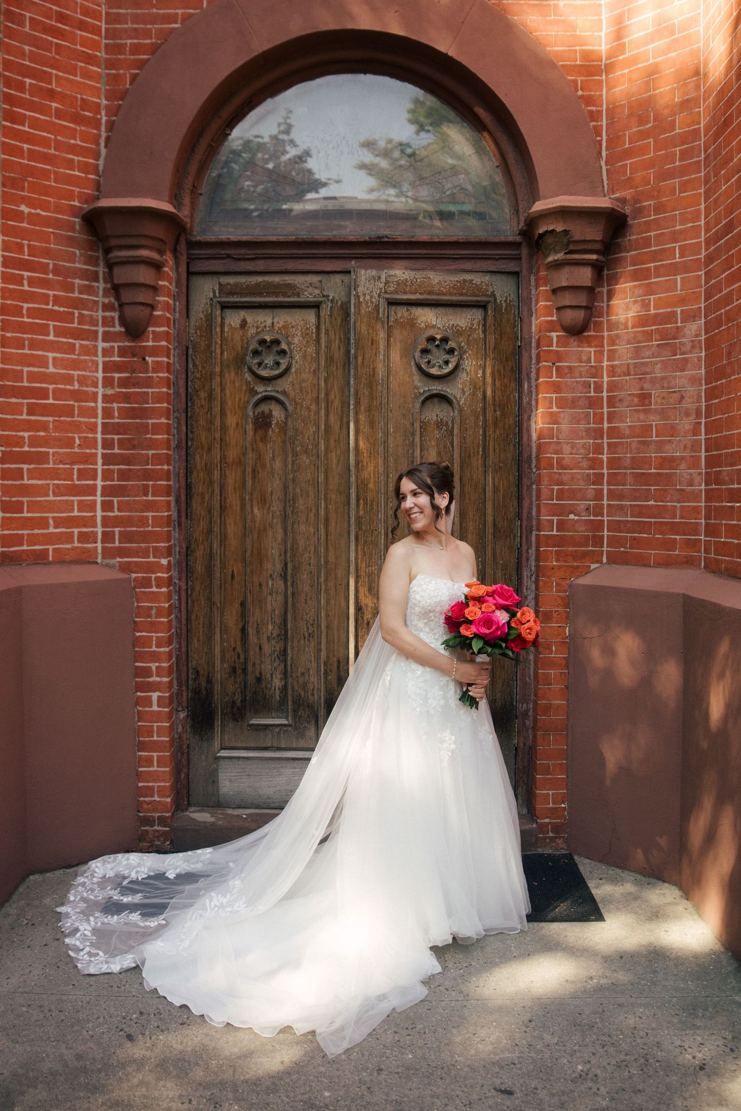 Bride in white strapless gown holding vibrant pink bouquet, posing before ornate wooden arched door on brick building.