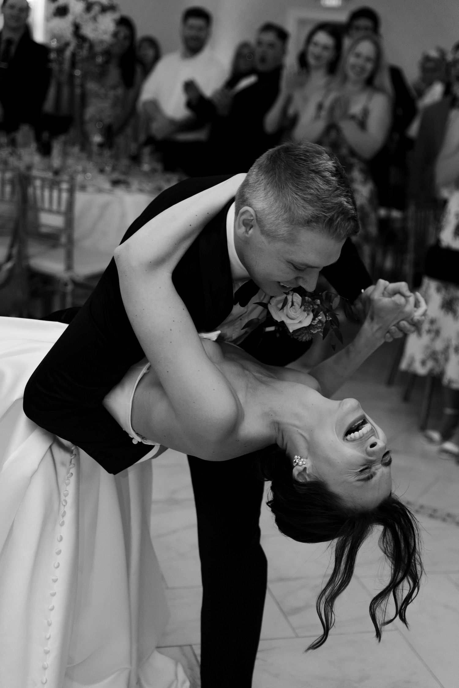 Black and white photo of groom dipping bride during first dance at wedding reception with guests applauding in background.
