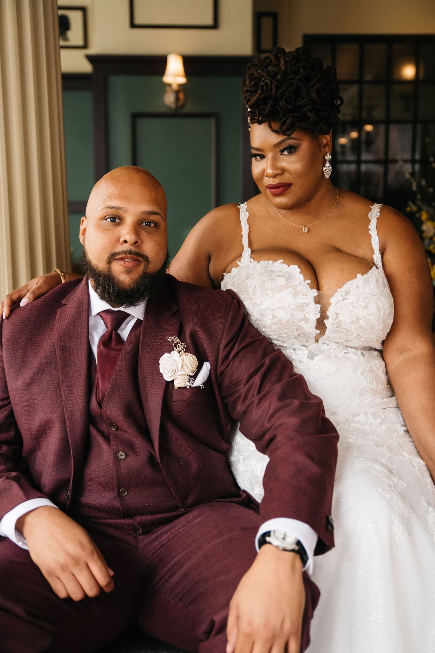 A bride and groom pose together, she in a white lace gown and he in a burgundy suit with boutonniere.