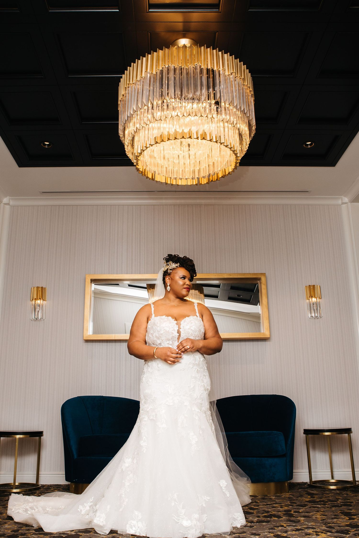 A bride in a white lace mermaid gown stands elegantly beneath a gold chandelier in a luxurious venue.