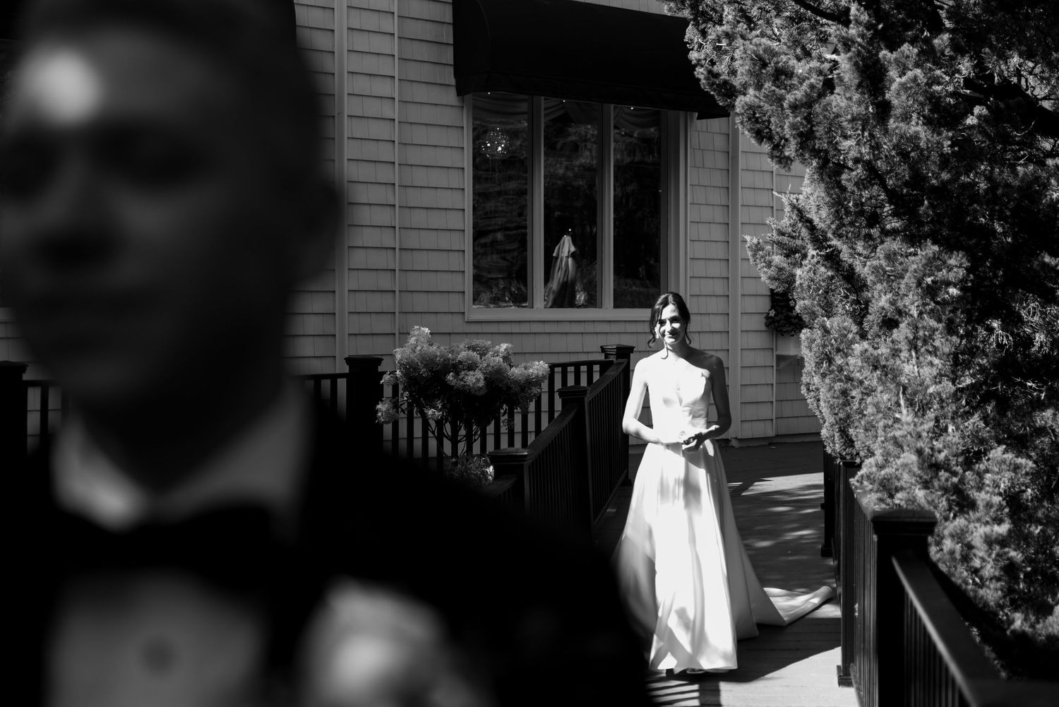 Black and white wedding photo of bride in elegant gown standing on wooden deck outside a house.