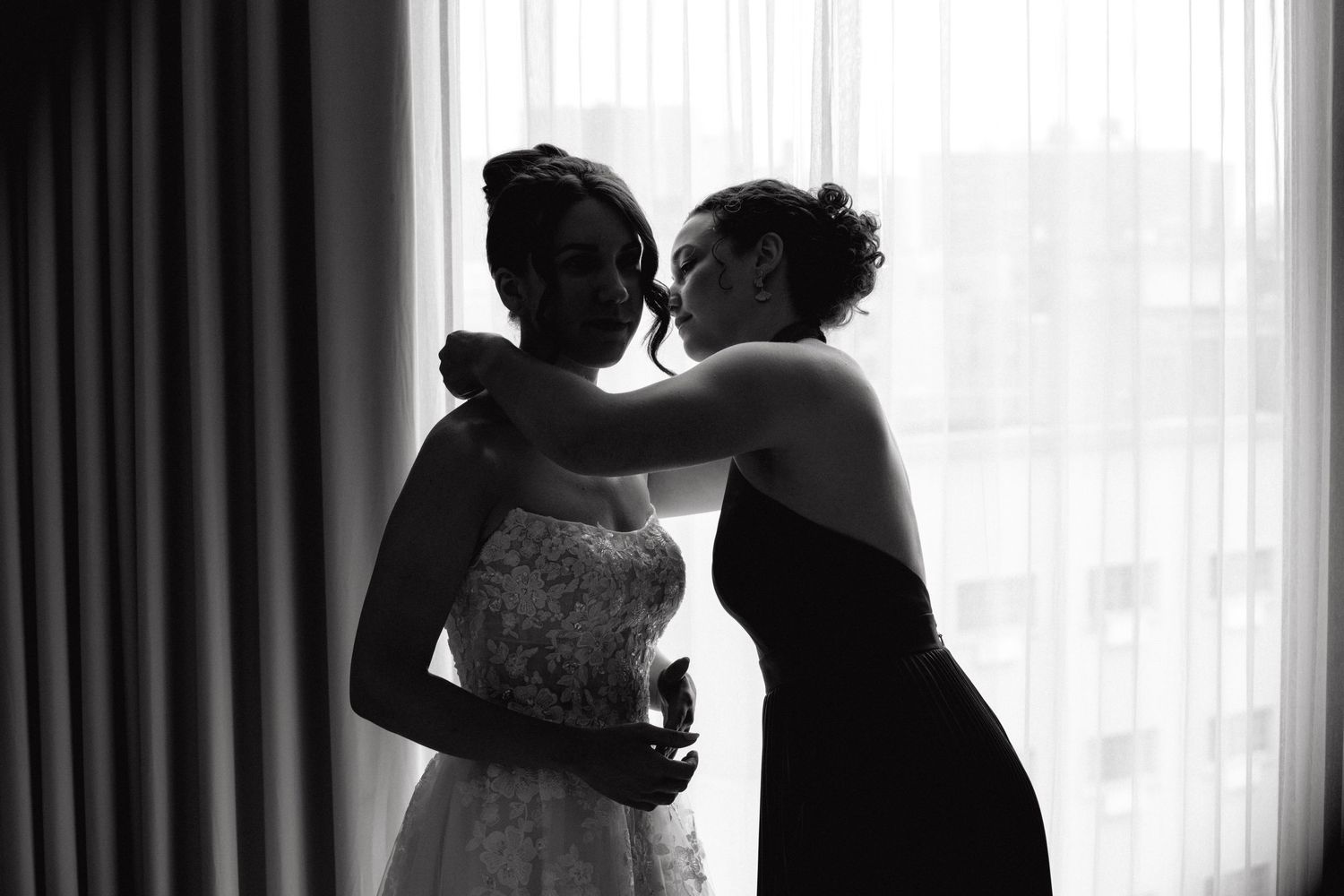 Black and white photo of a bridesmaid helping a bride in a lace wedding dress near a bright window.