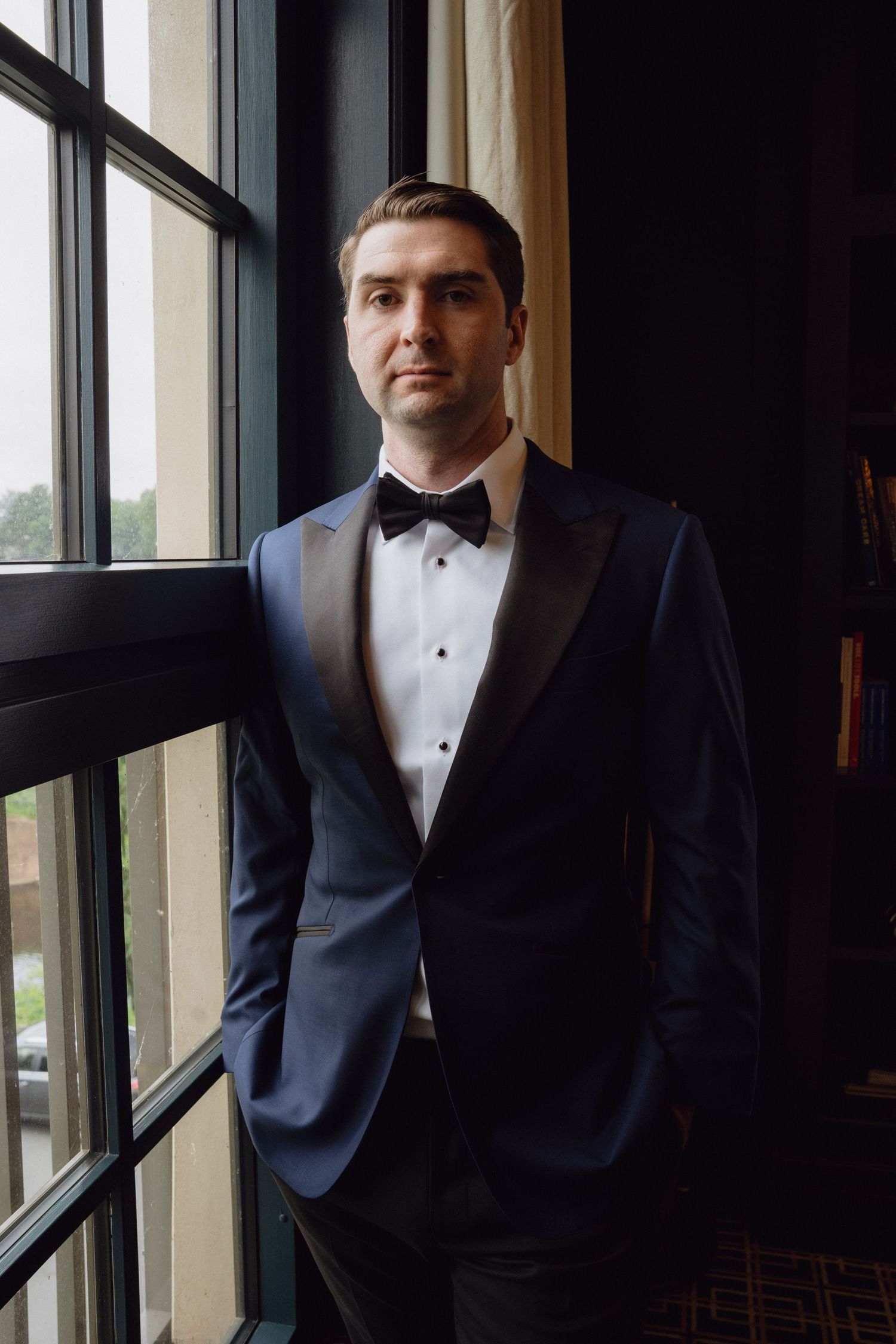 A man in a navy blue tuxedo with black bow tie stands confidently by large windows in elegant indoor setting.
