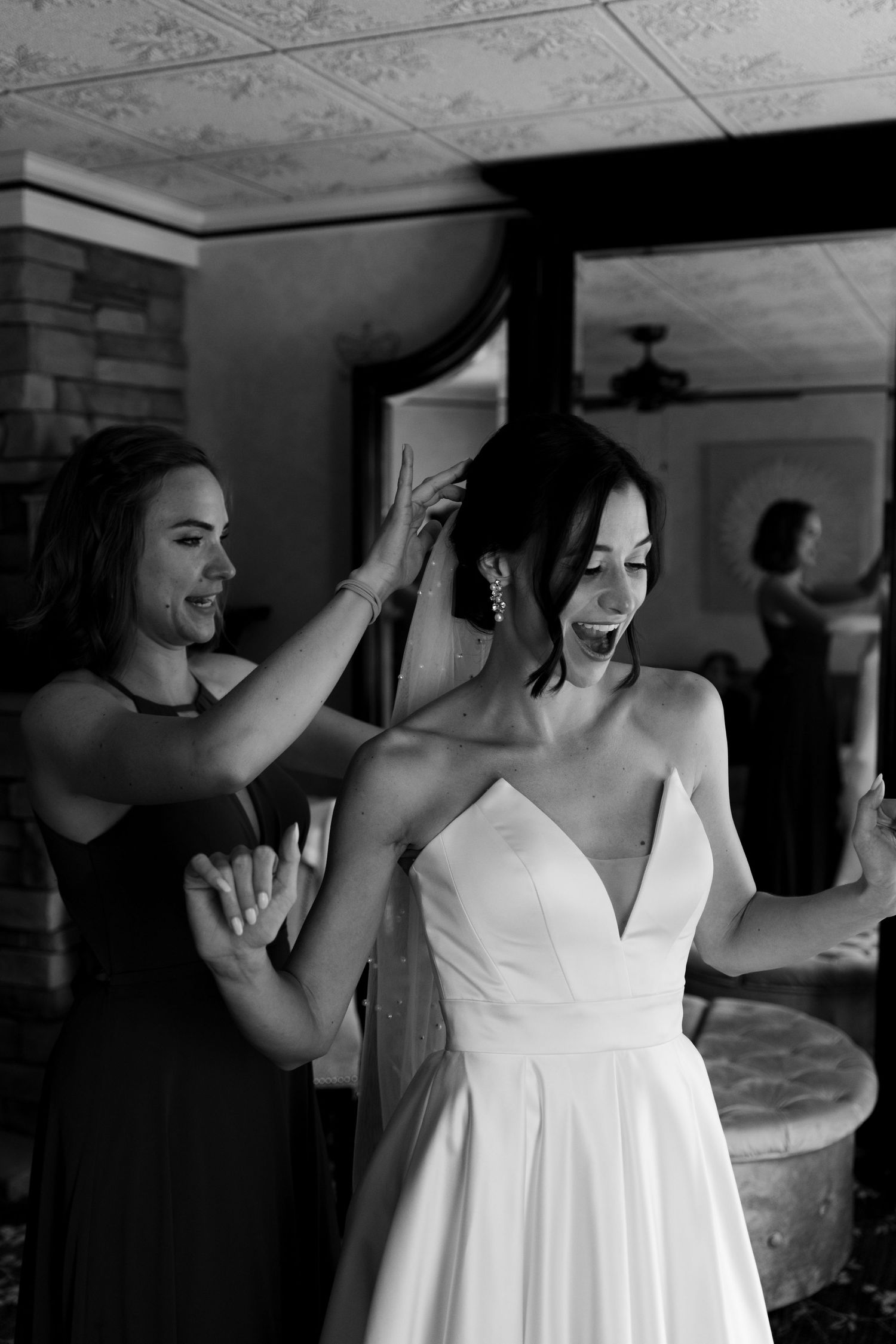 Black and white photo of a bride in a white gown smiling as her bridesmaid adjusts her veil indoors.
