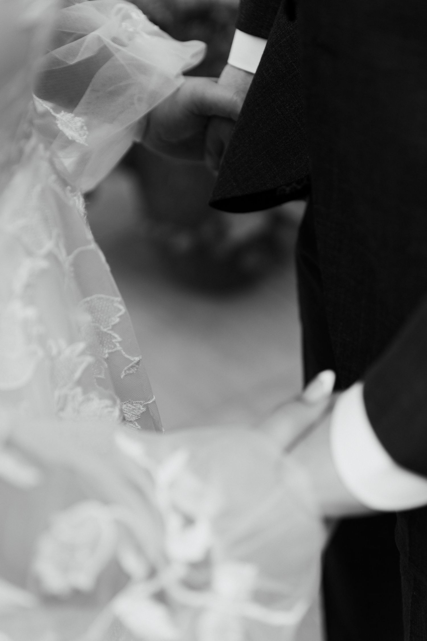 Black and white wedding photo showing bride in lace gown and veil holding hands with groom in dark suit.
