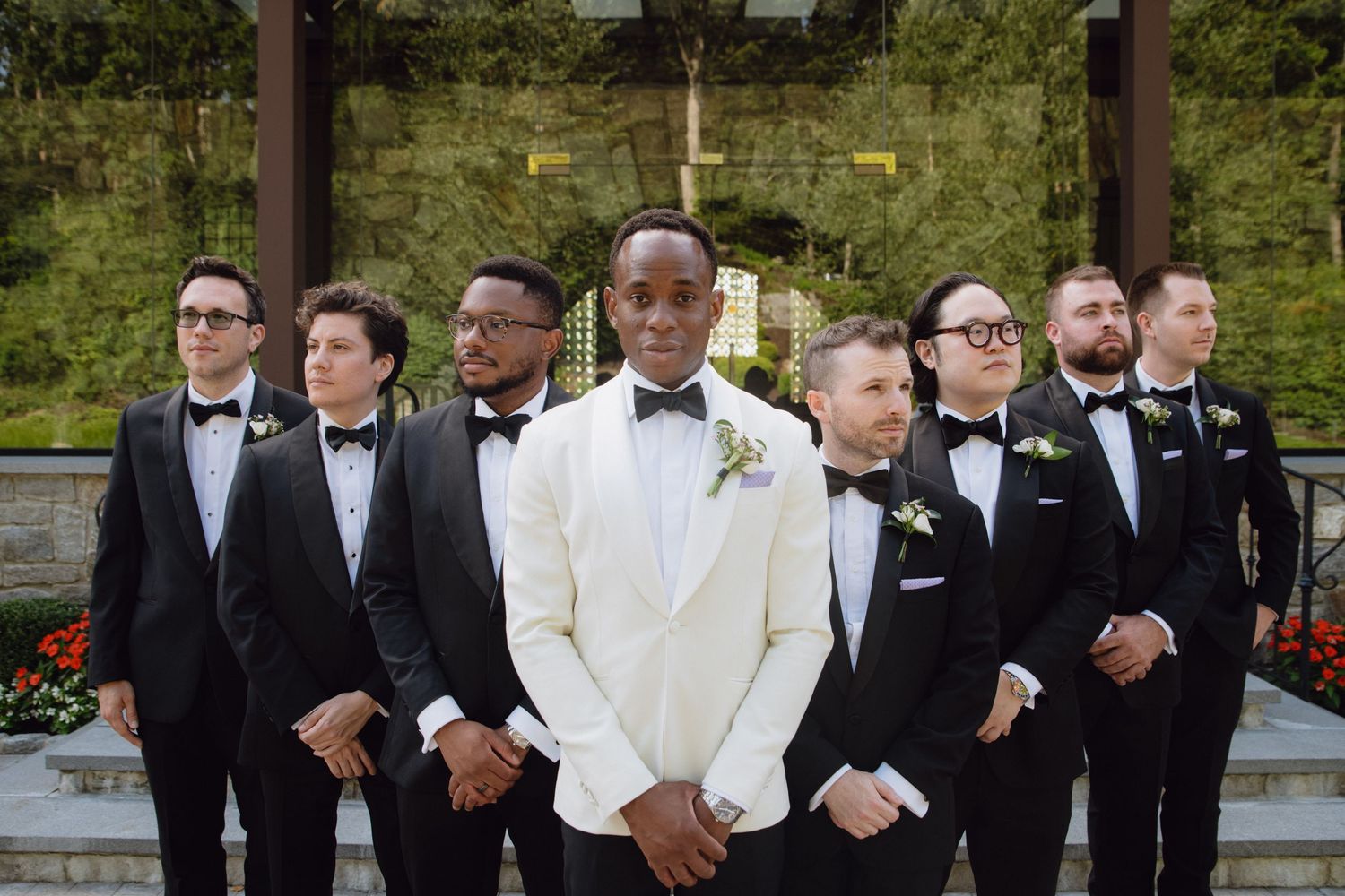 Groom in white tuxedo jacket stands with groomsmen in black tuxedos outside a chapel wedding venue.