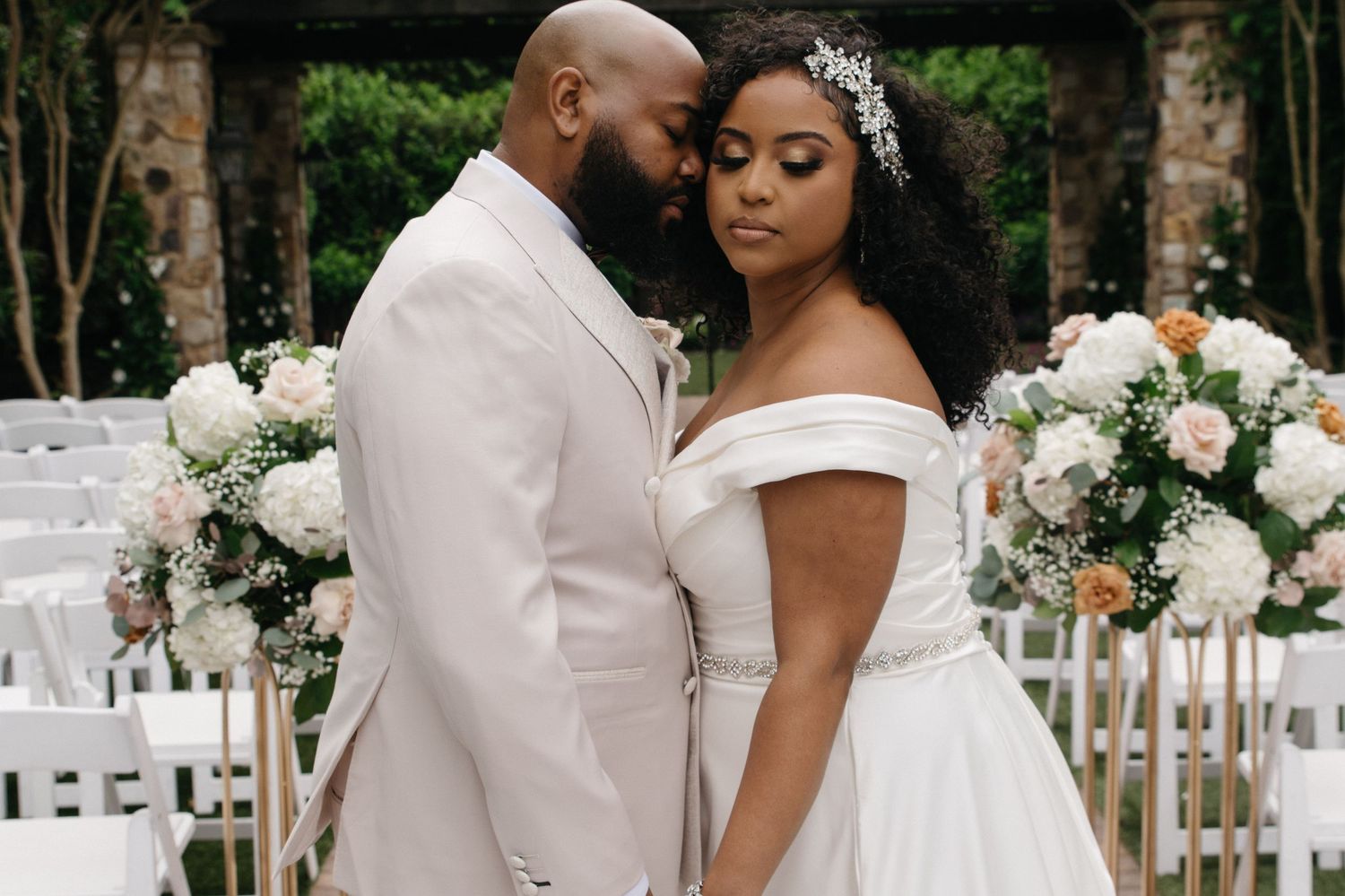 Elegant bride and groom pose together at outdoor wedding ceremony with white chairs and floral arrangements.