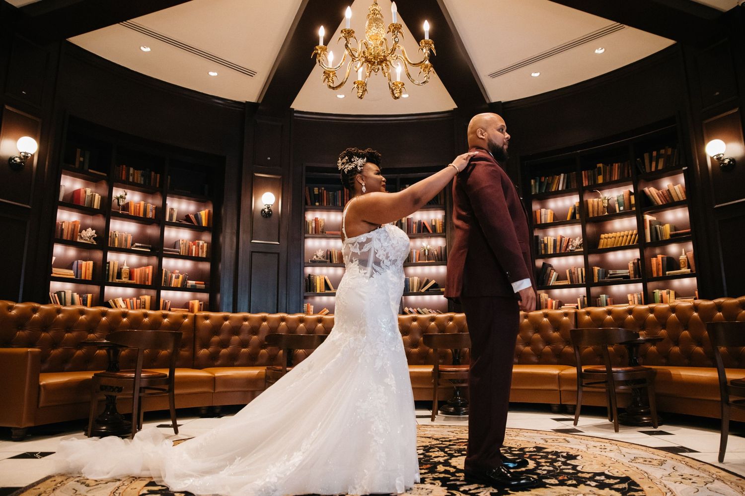 Bride in white gown adjusts groom's burgundy suit in elegant library venue with chandelier and leather sofas.