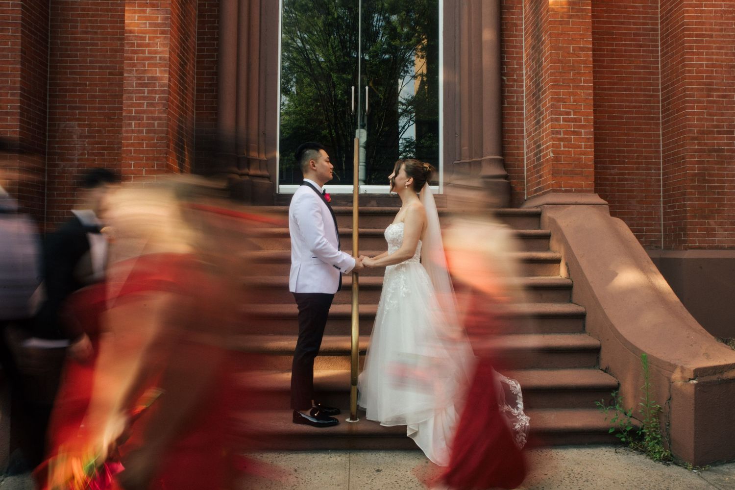 Bride and groom hold hands on church steps while blurred wedding guests in red pass by in foreground.