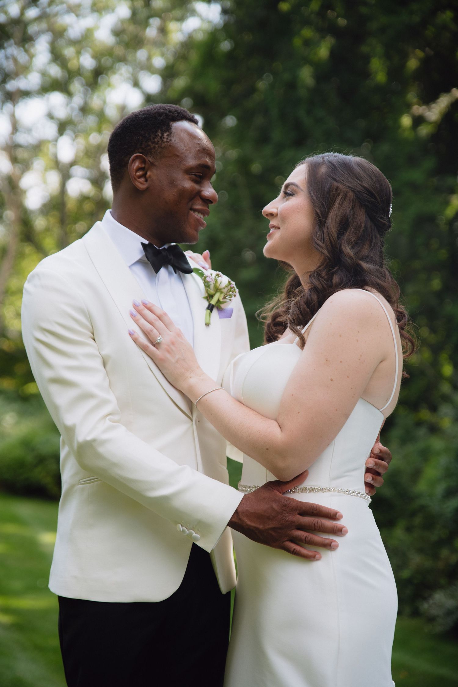 Interracial couple in wedding attire embracing outdoors, groom in white tuxedo, bride in white gown, smiling at each other.