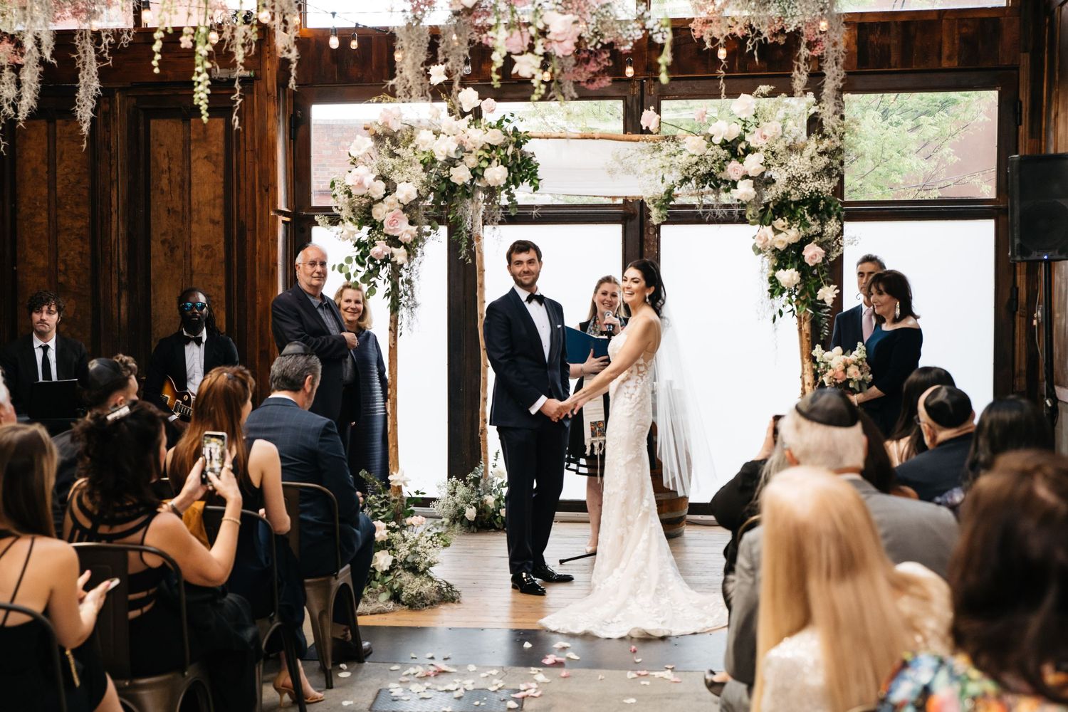 Couple exchanges vows at elegant indoor wedding ceremony with floral arch, surrounded by seated guests in a rustic barn venue.