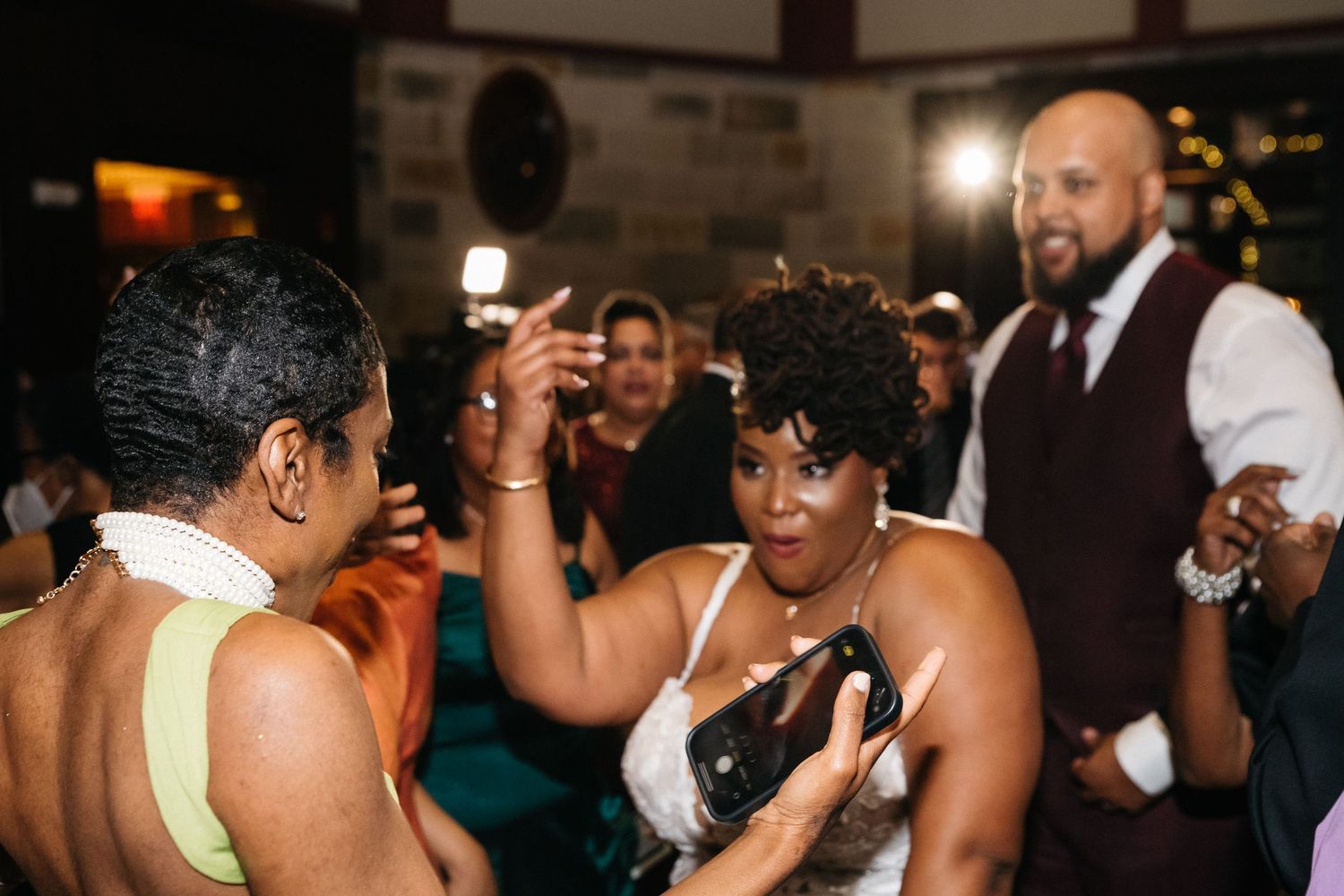 Guests dancing joyfully at a wedding reception, with a bearded man in a vest visible in the background.