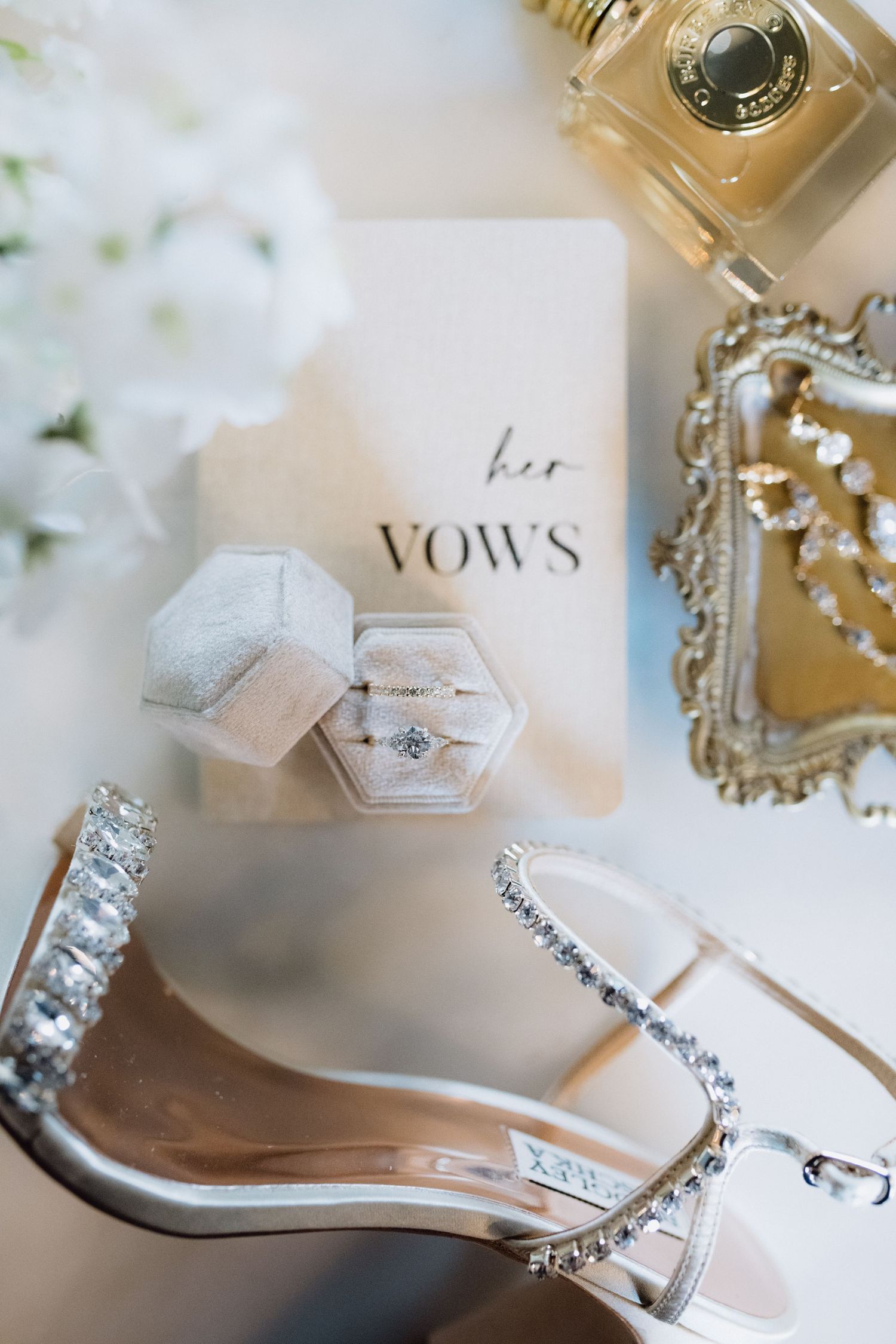 Elegant bridal flat lay featuring diamond heels, ring box with engagement ring, vow book, perfume bottle, and crystal jewelry on white.