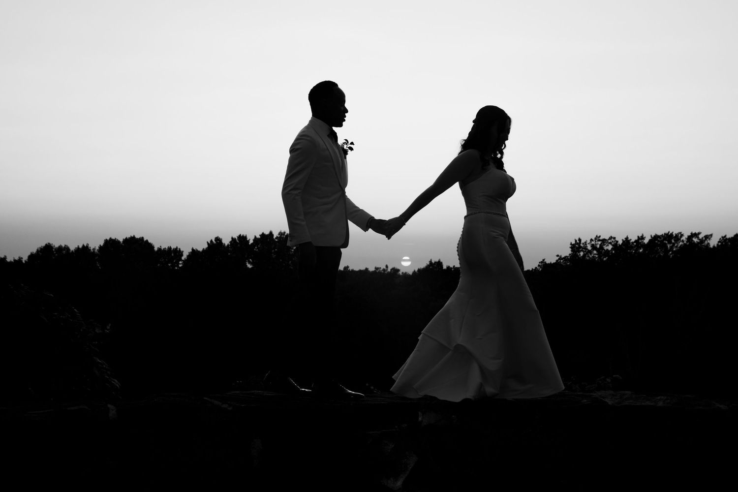 Black and white silhouette of a bride and groom holding hands at sunset with trees in the background.