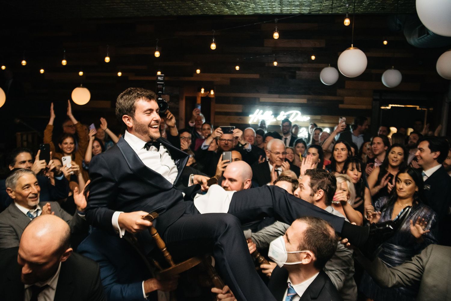Groom lifted by groomsmen during lively wedding reception celebration with string lights and cheering guests.