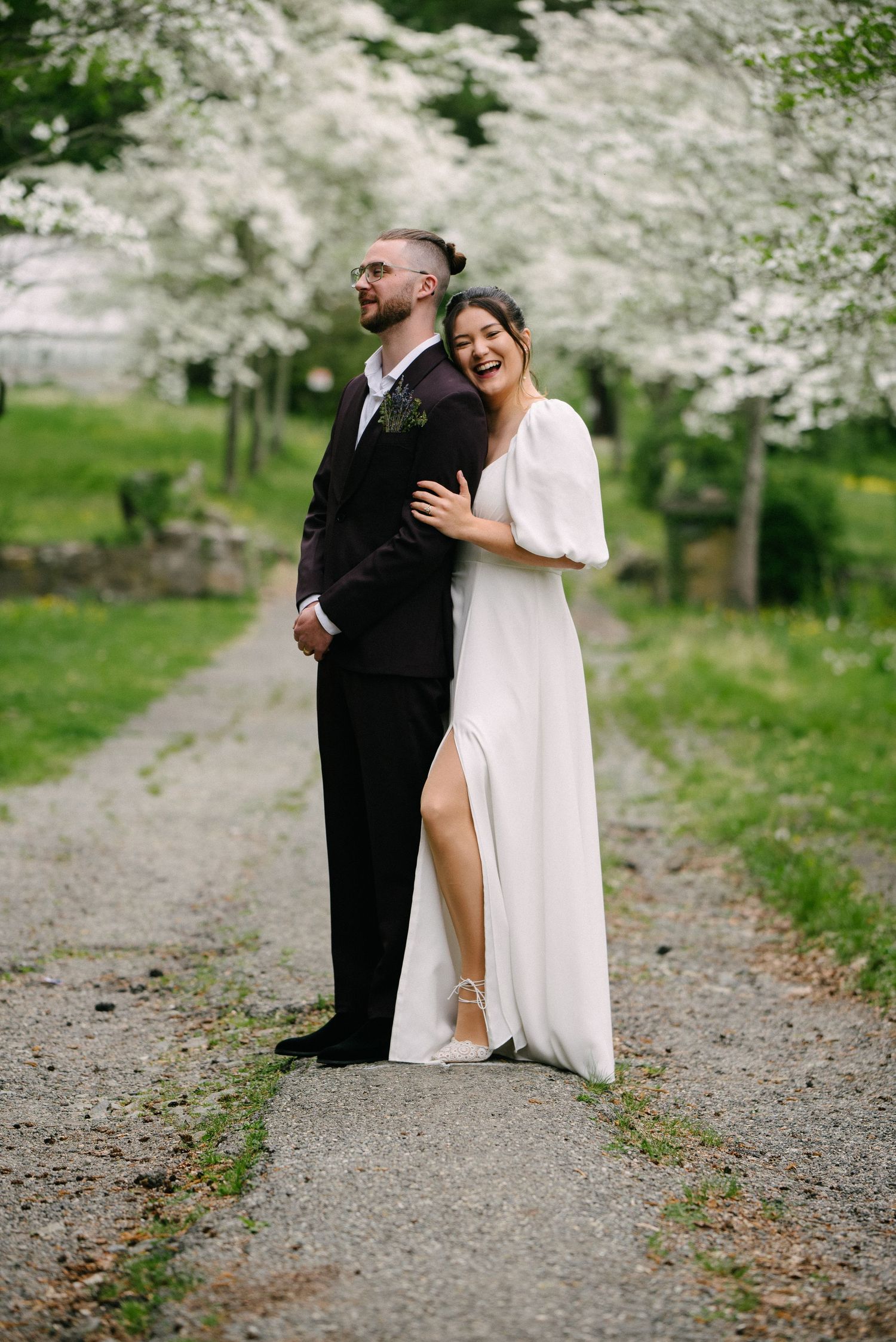 Bride in white gown laughs hugging groom in dark suit on a tree-lined path with blooming white trees.