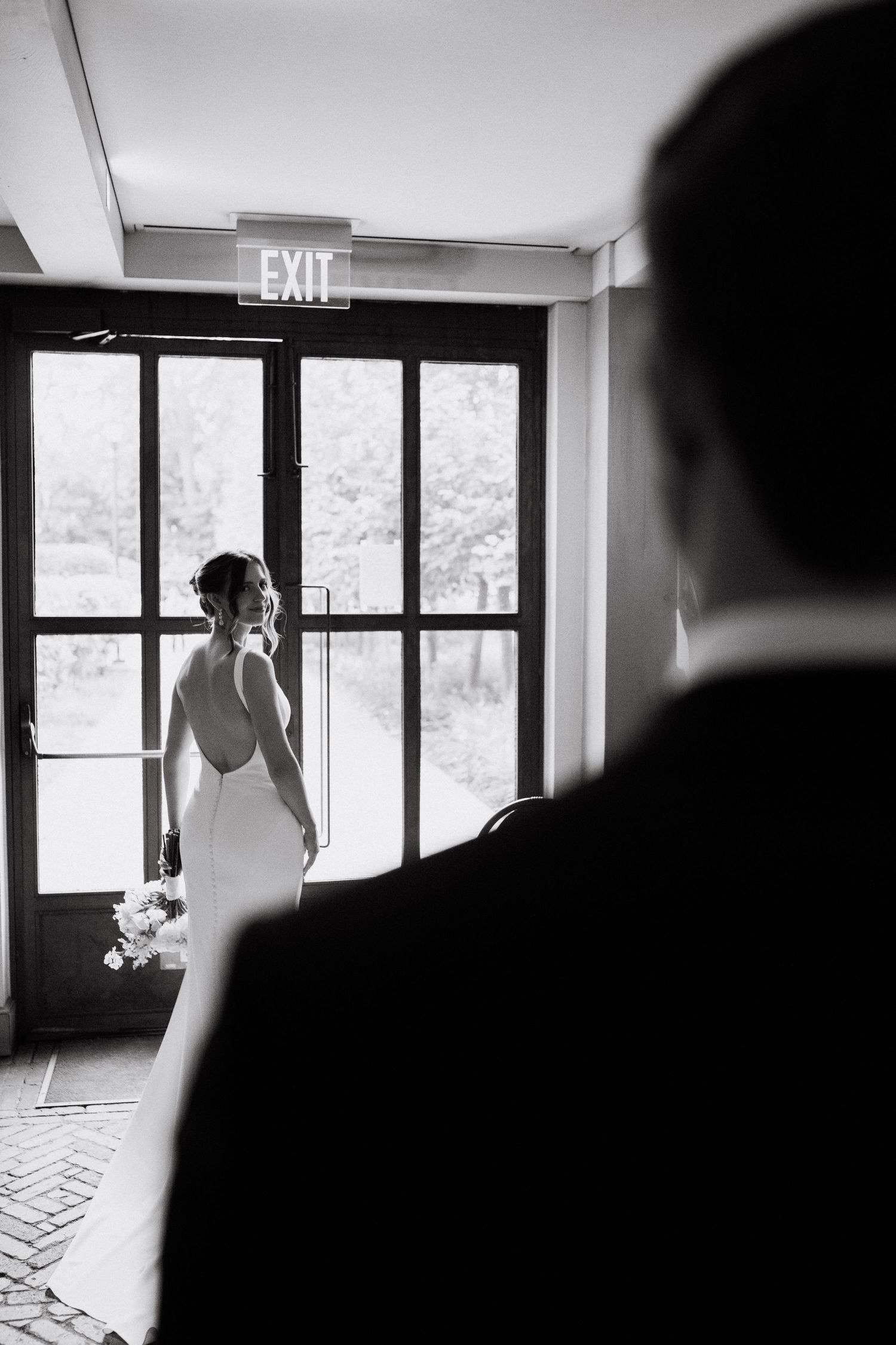 Black and white wedding photo of bride in backless gown near exit door as groom watches from foreground.