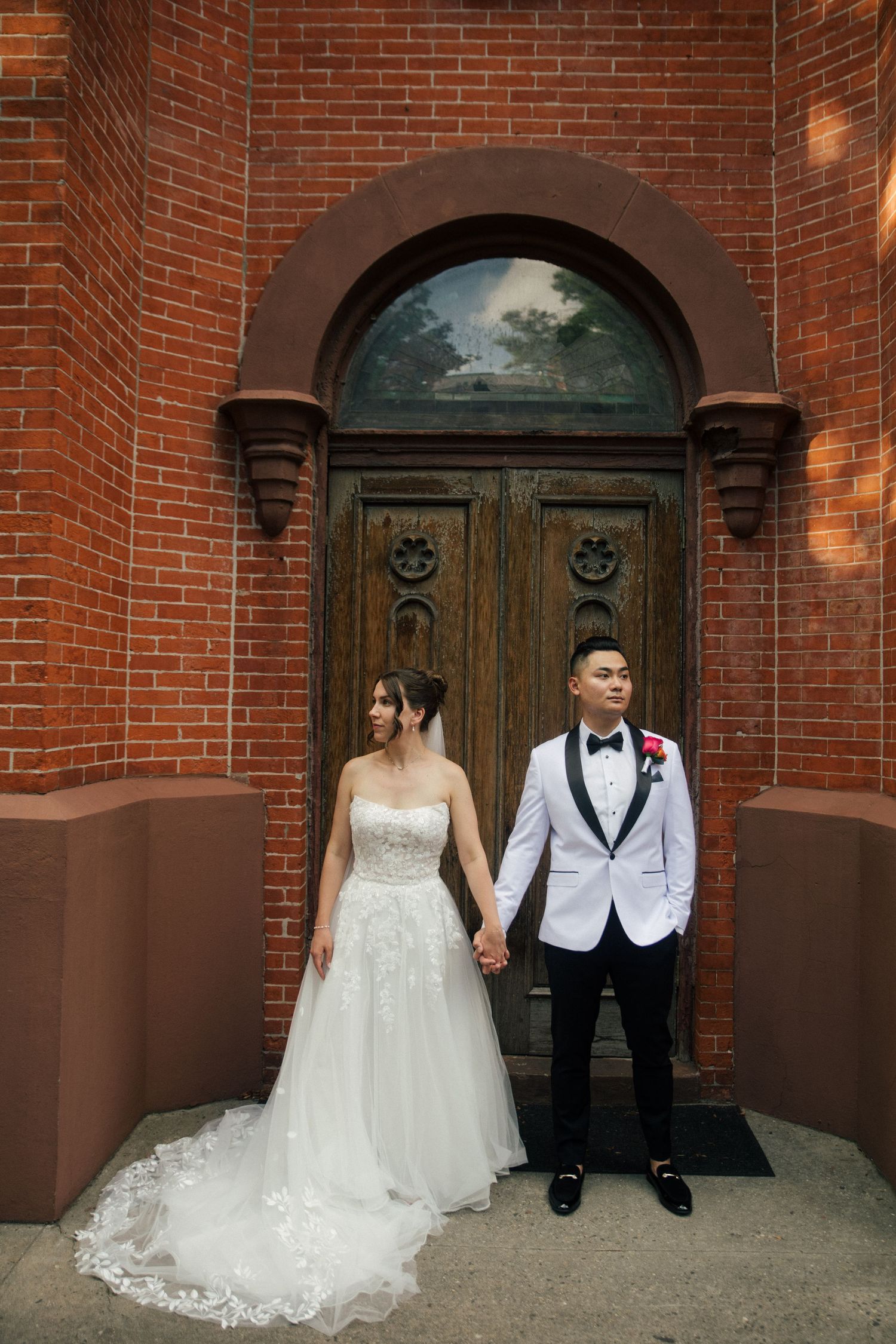 Bride and groom holding hands in front of ornate wooden door and red brick church wall on their wedding day.