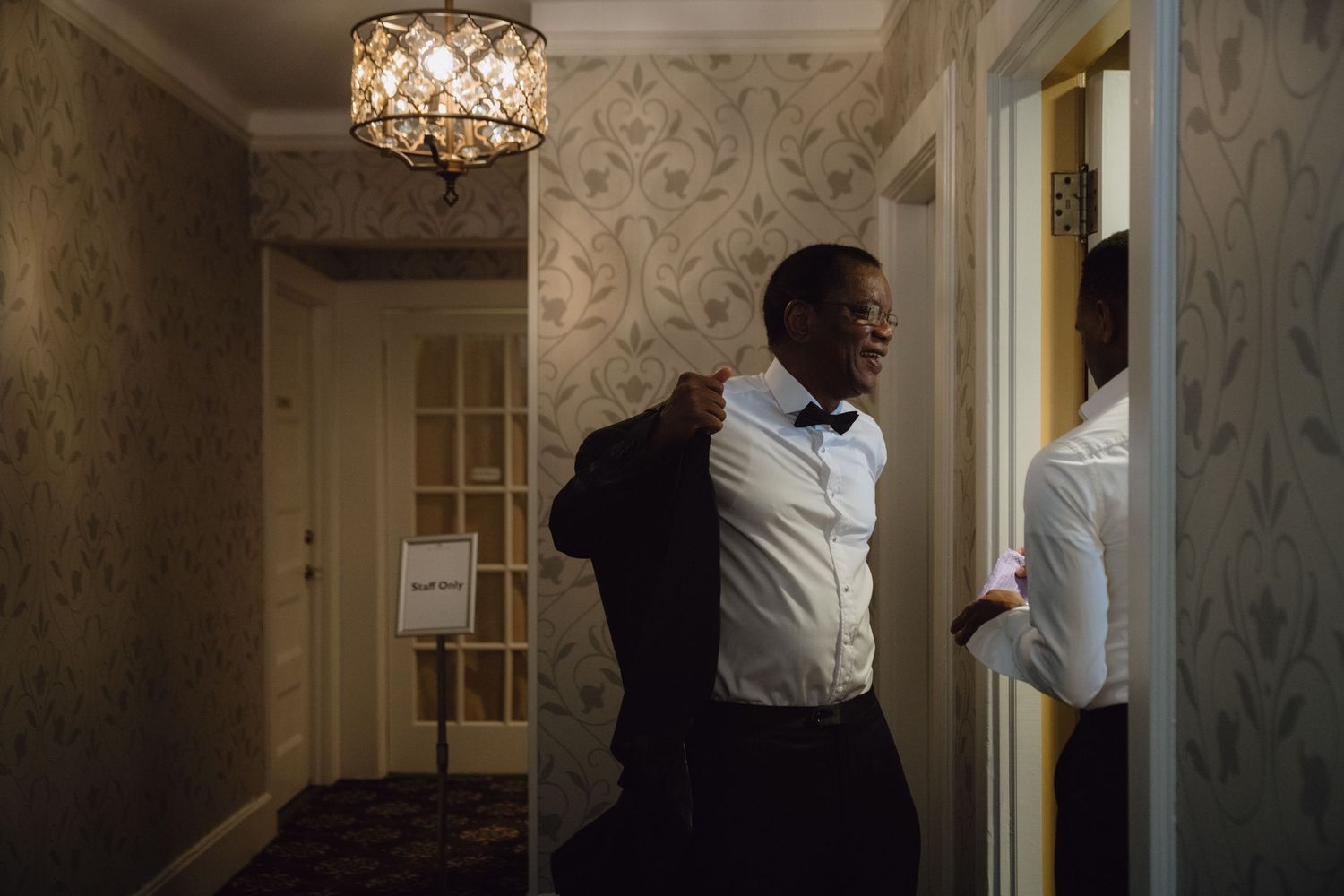 A groom in a tuxedo and bow tie puts on his jacket while checking his reflection in a mirror before his wedding.
