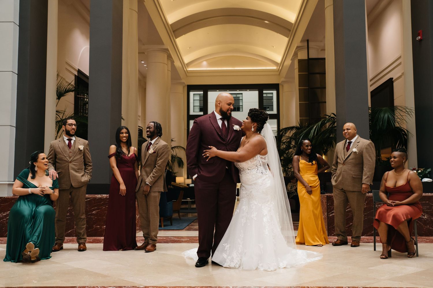 Bride and groom share first dance surrounded by wedding party in elegant venue with arched ceilings.