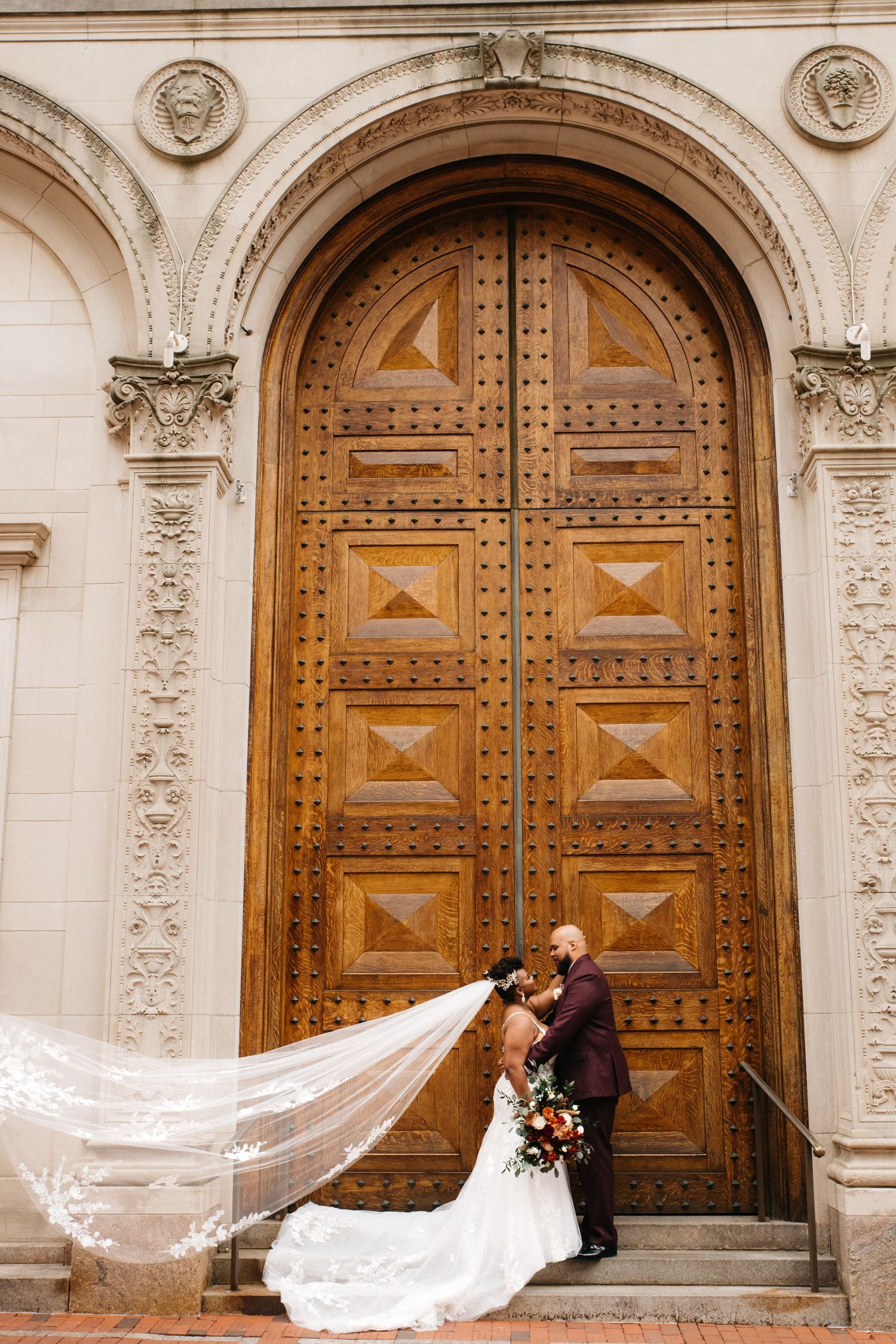 Bride and groom kissing in front of ornate wooden arched doors, with veil flowing dramatically in the wind.