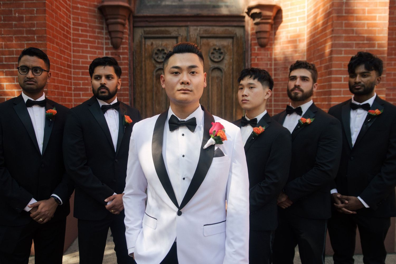 Groom in white tuxedo with groomsmen in black suits posing outdoors before a brick building wedding photo.