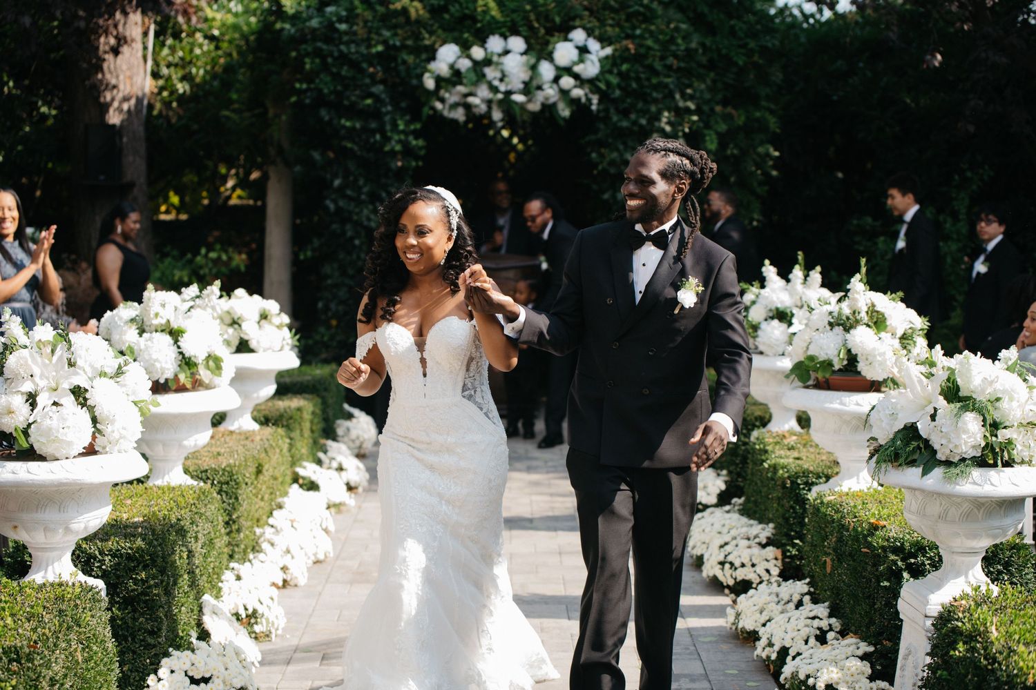 Joyful newlyweds walk down floral aisle lined with white hydrangeas in urns after outdoor wedding ceremony.