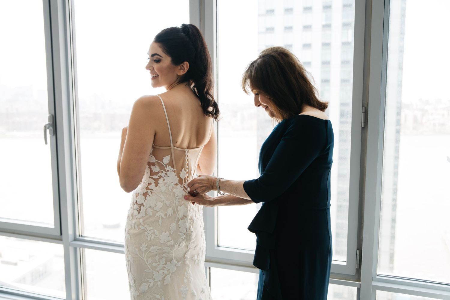 Bride in lace wedding gown being helped by woman in black suit near large windows with city view.