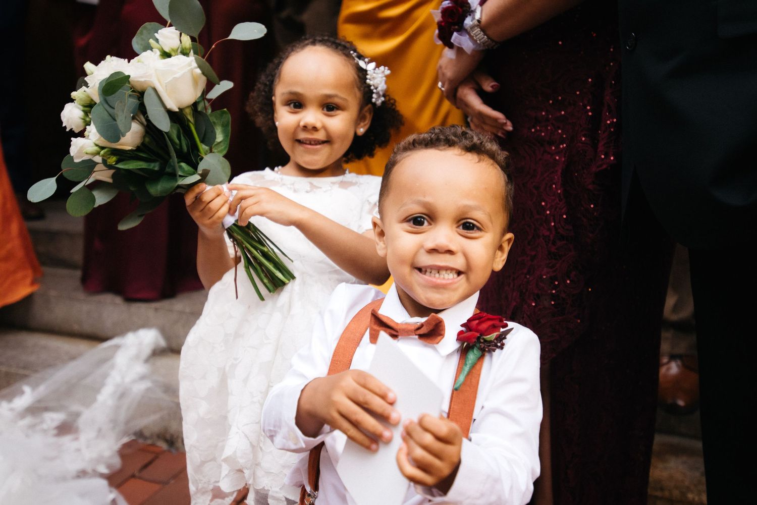 Adorable flower girl holding white bouquet and ring bearer in suspenders smiling at a wedding ceremony.