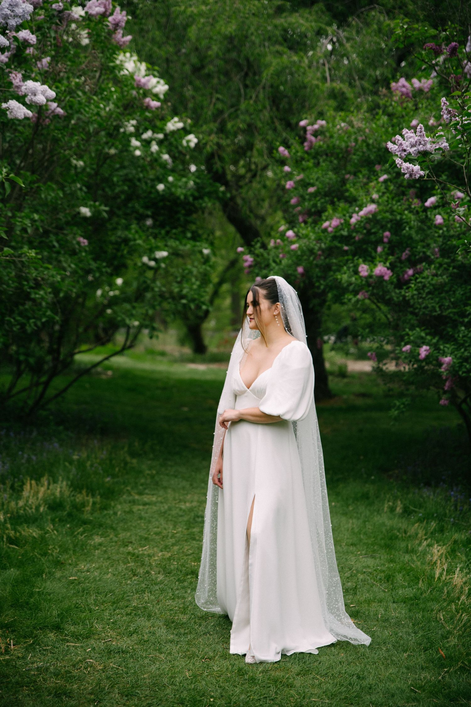 Elegant bride in white flowing gown and veil standing among blooming lilac trees in lush green garden.