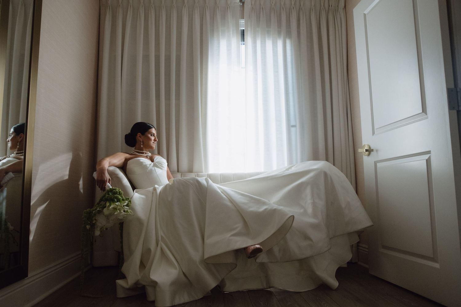 Bride in white gown resting on chaise lounge by curtained window, holding bouquet, in soft natural light.