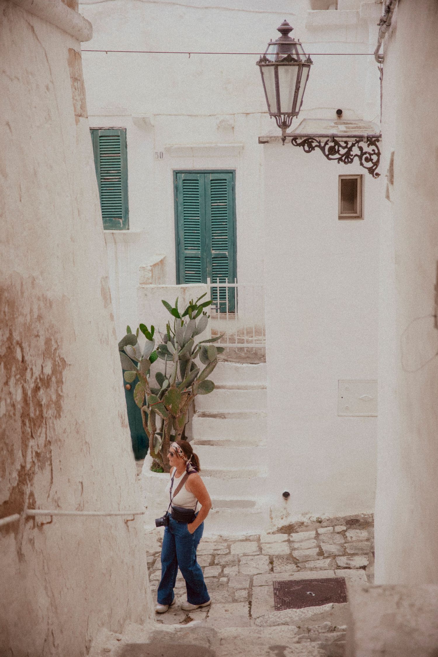 Person standing in a charming Mediterranean alley with green shutters and vintage lantern.