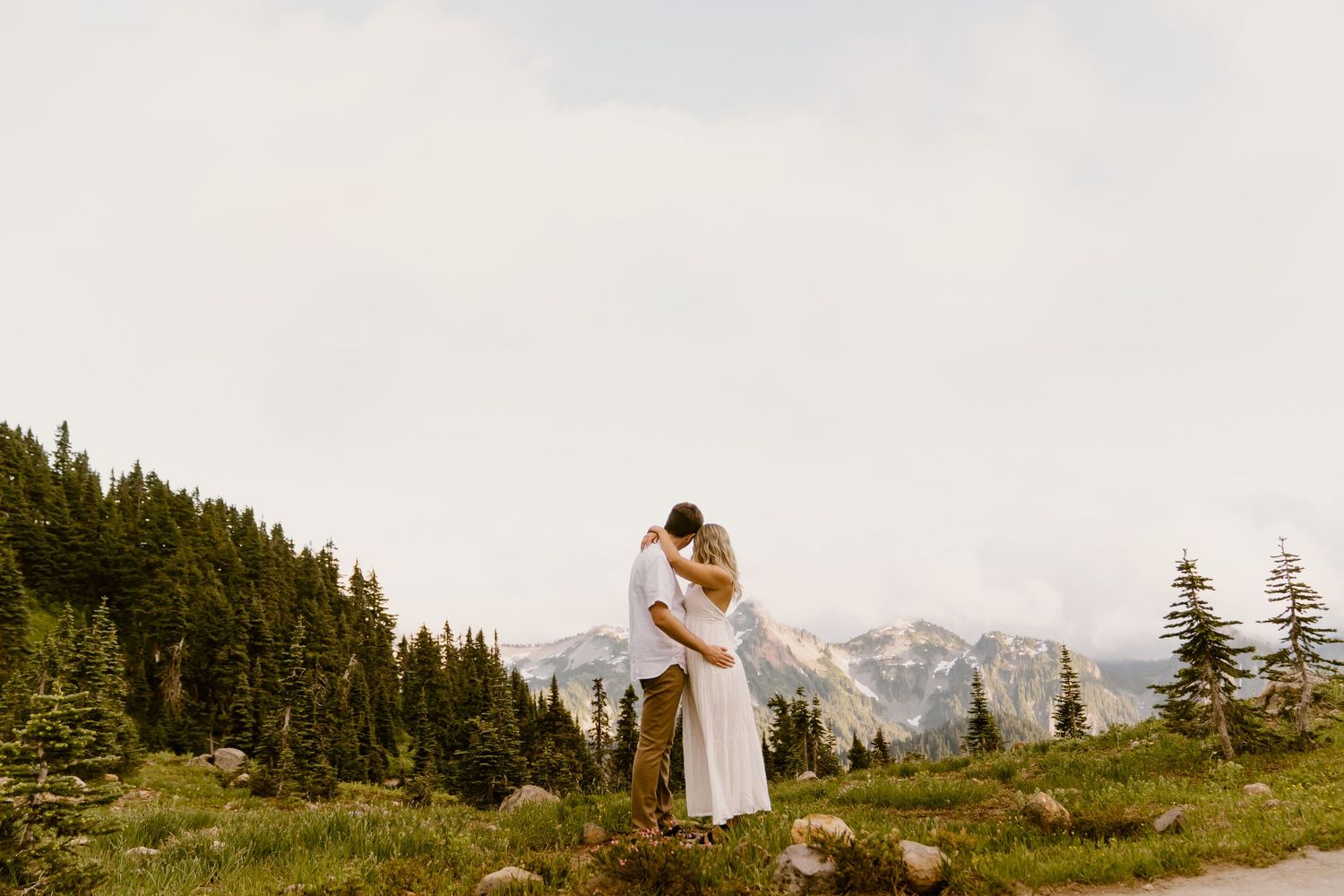 Couple embracing and kissing in a mountain meadow surrounded by evergreen trees and misty peaks.