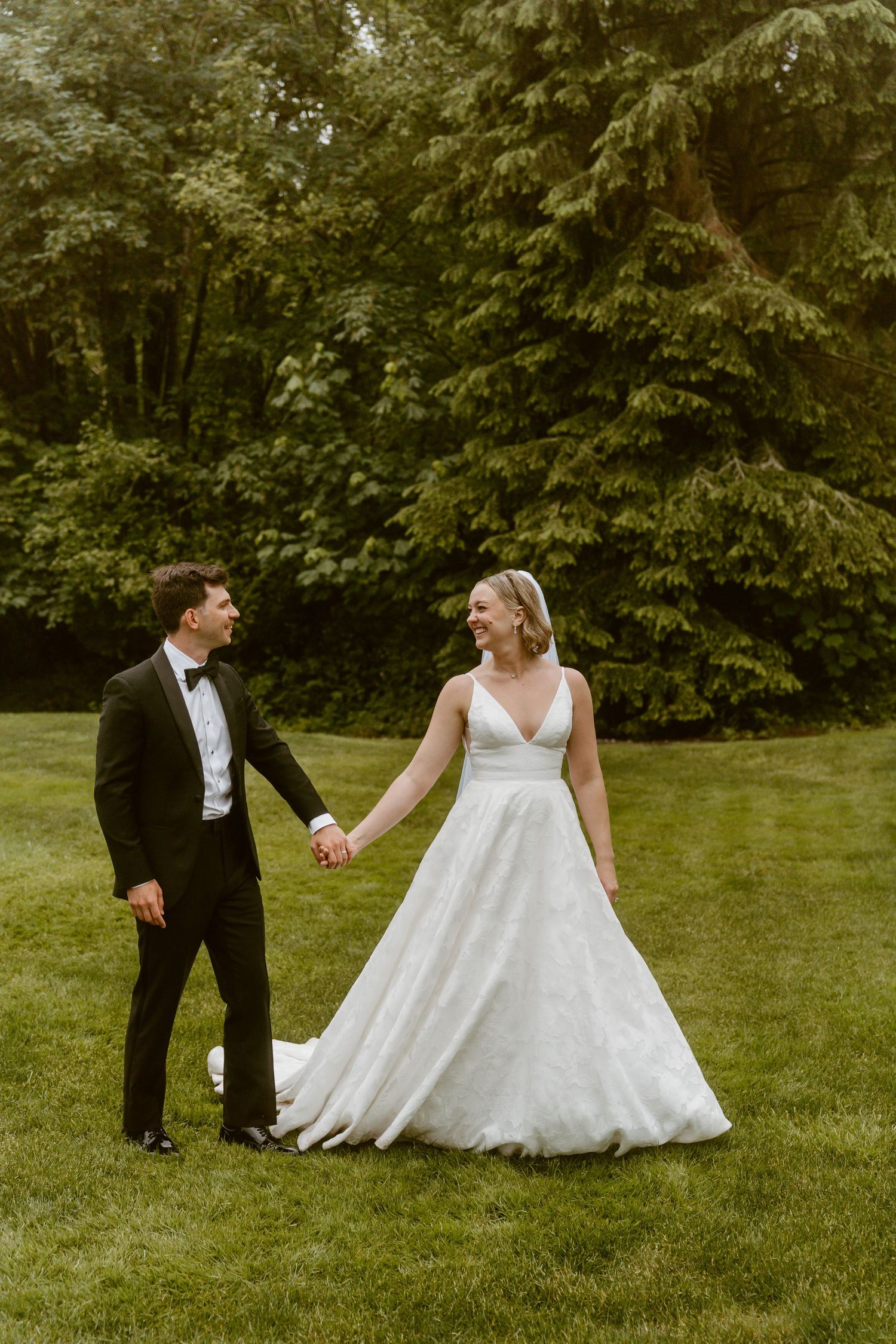 Smiling bride and groom holding hands while walking on green lawn surrounded by tall trees.
