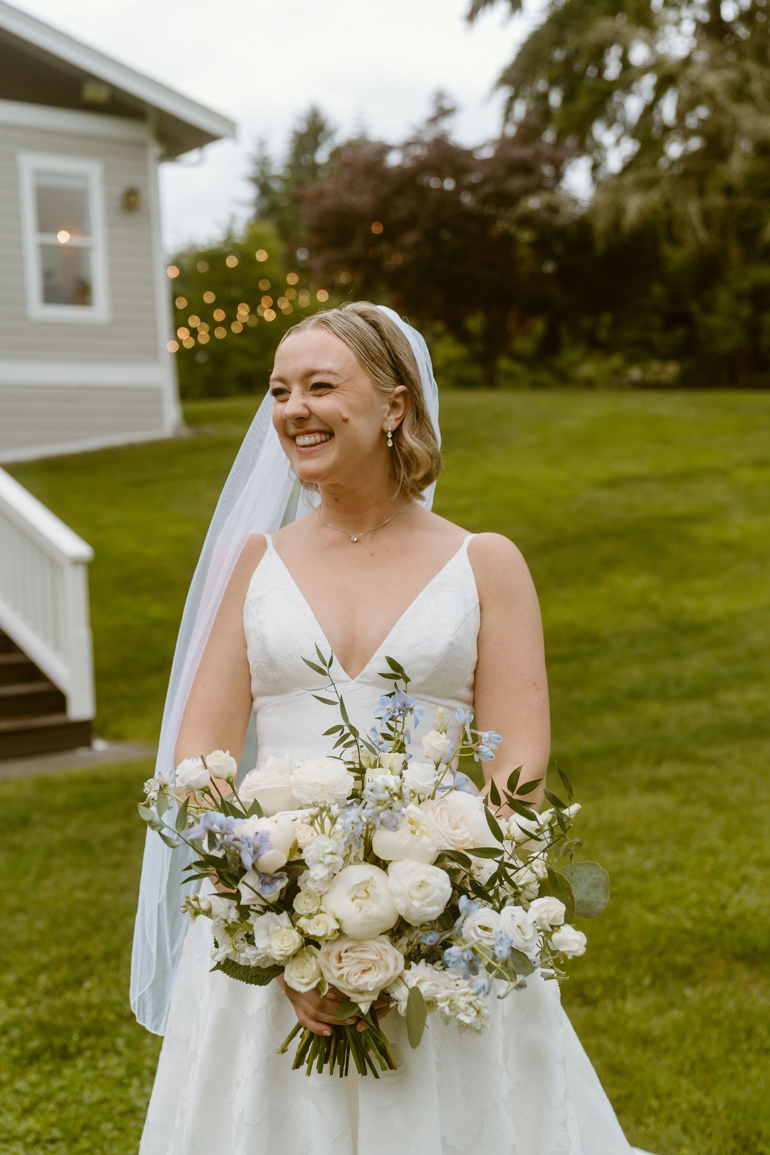 Radiant bride in white A-line gown holding white and blue bouquet laughing outdoors at venue.