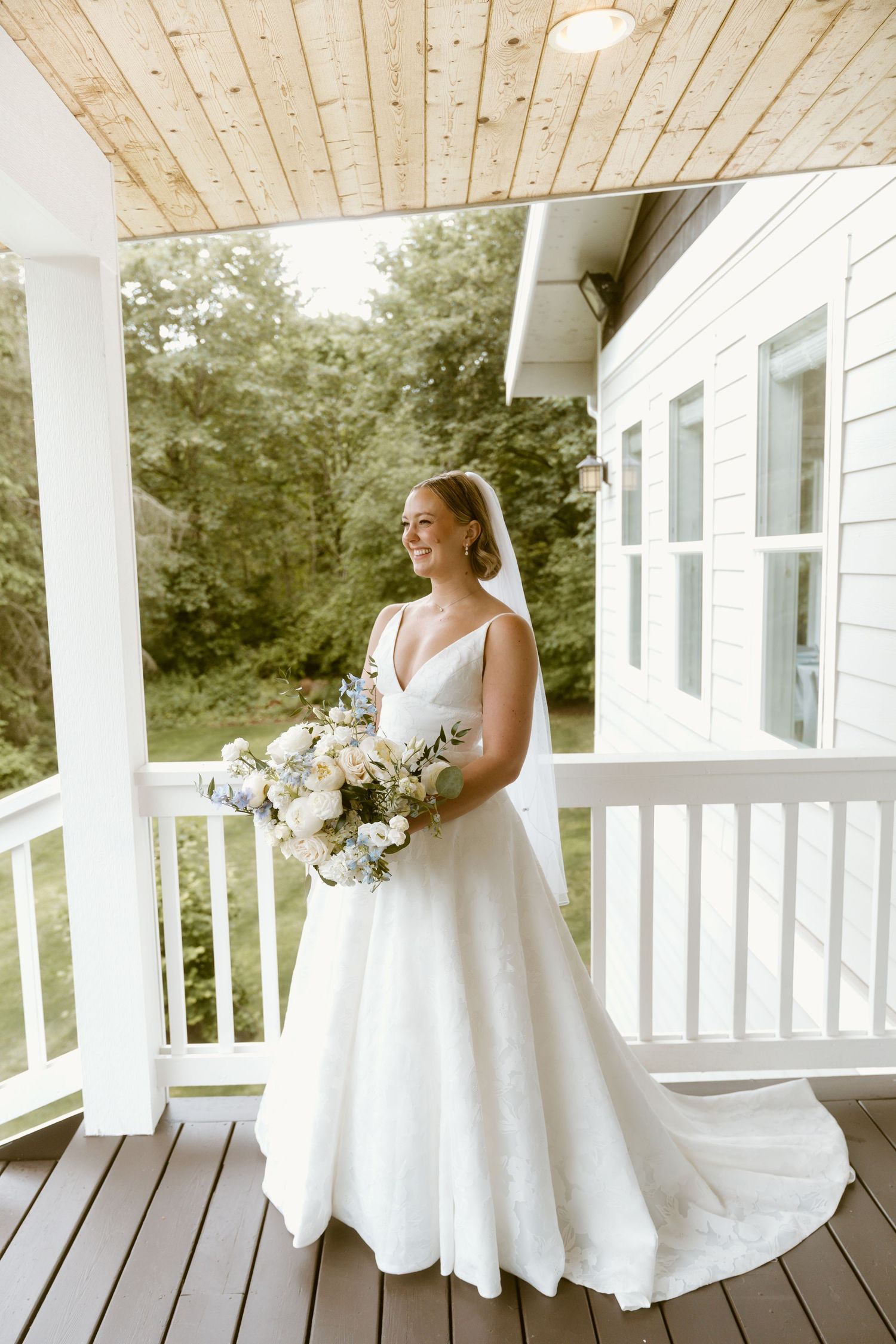 Bride in white gown holding floral bouquet smiling on a white wooden porch.