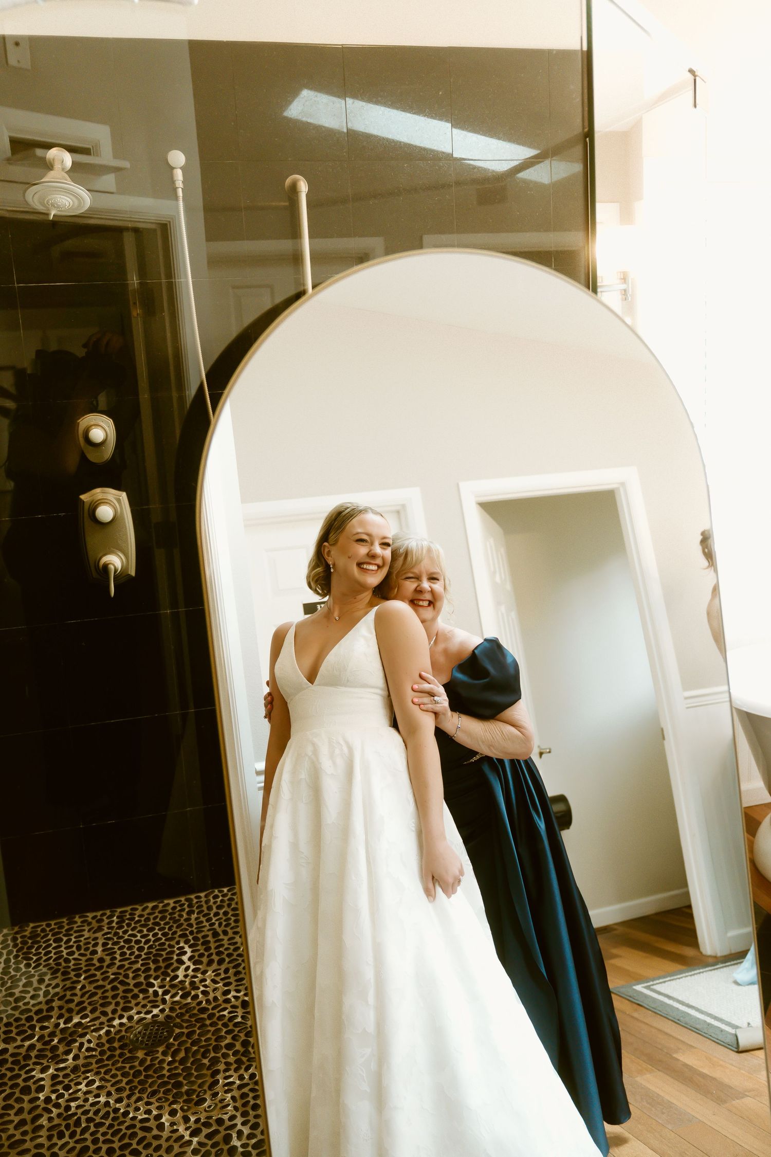 Bride in white gown smiling at mirror while woman in navy dress helps her prepare for wedding day.