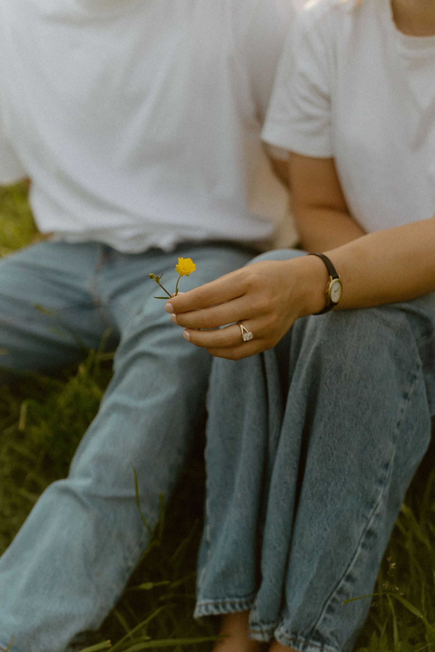 Couple sitting on grass in white tees and jeans, one holding a small yellow wildflower, wearing a ring and bracelet.