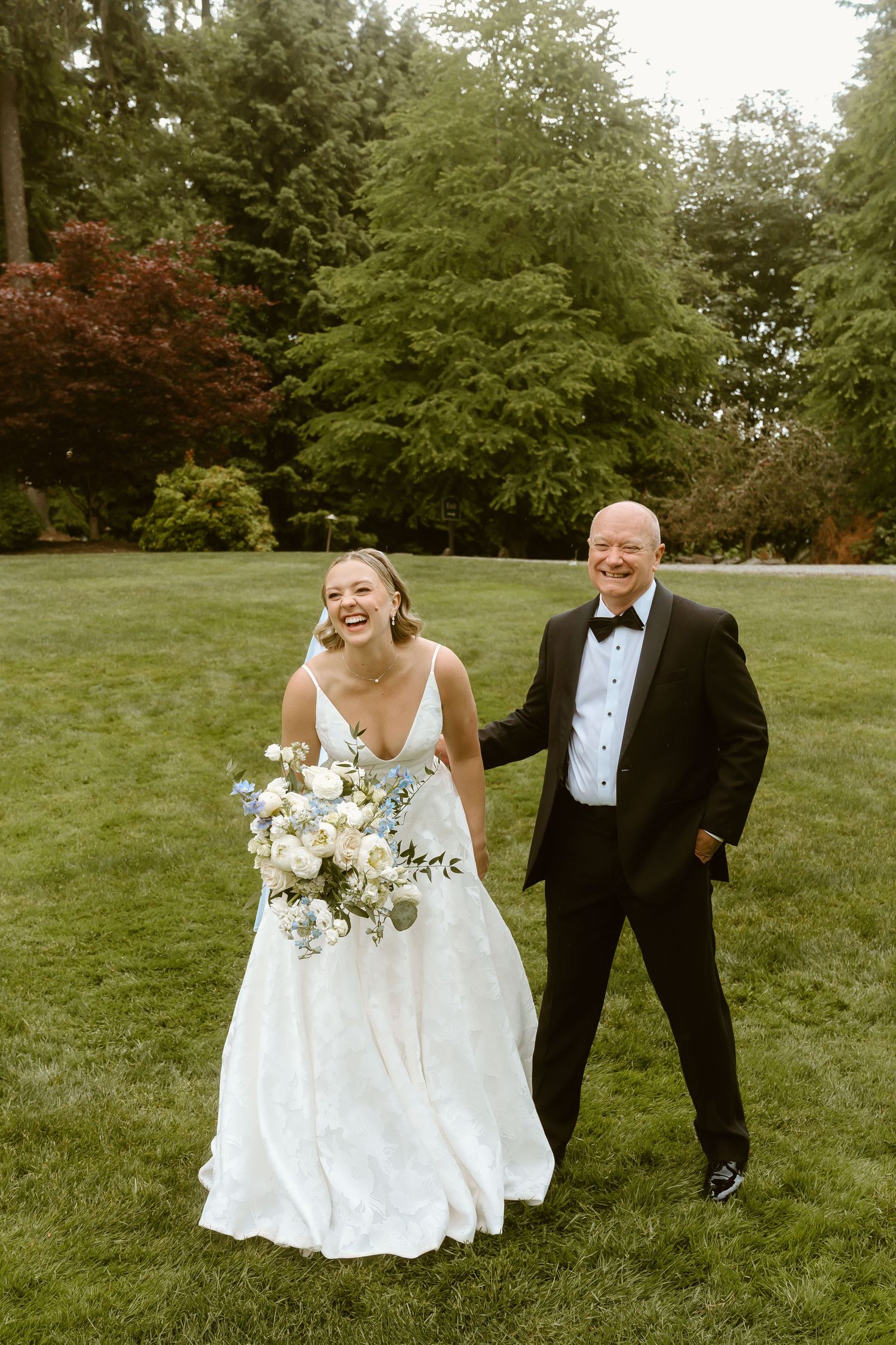 Joyful bride in white gown holding bouquet walks with older gentleman in tuxedo on green lawn at outdoor wedding.
